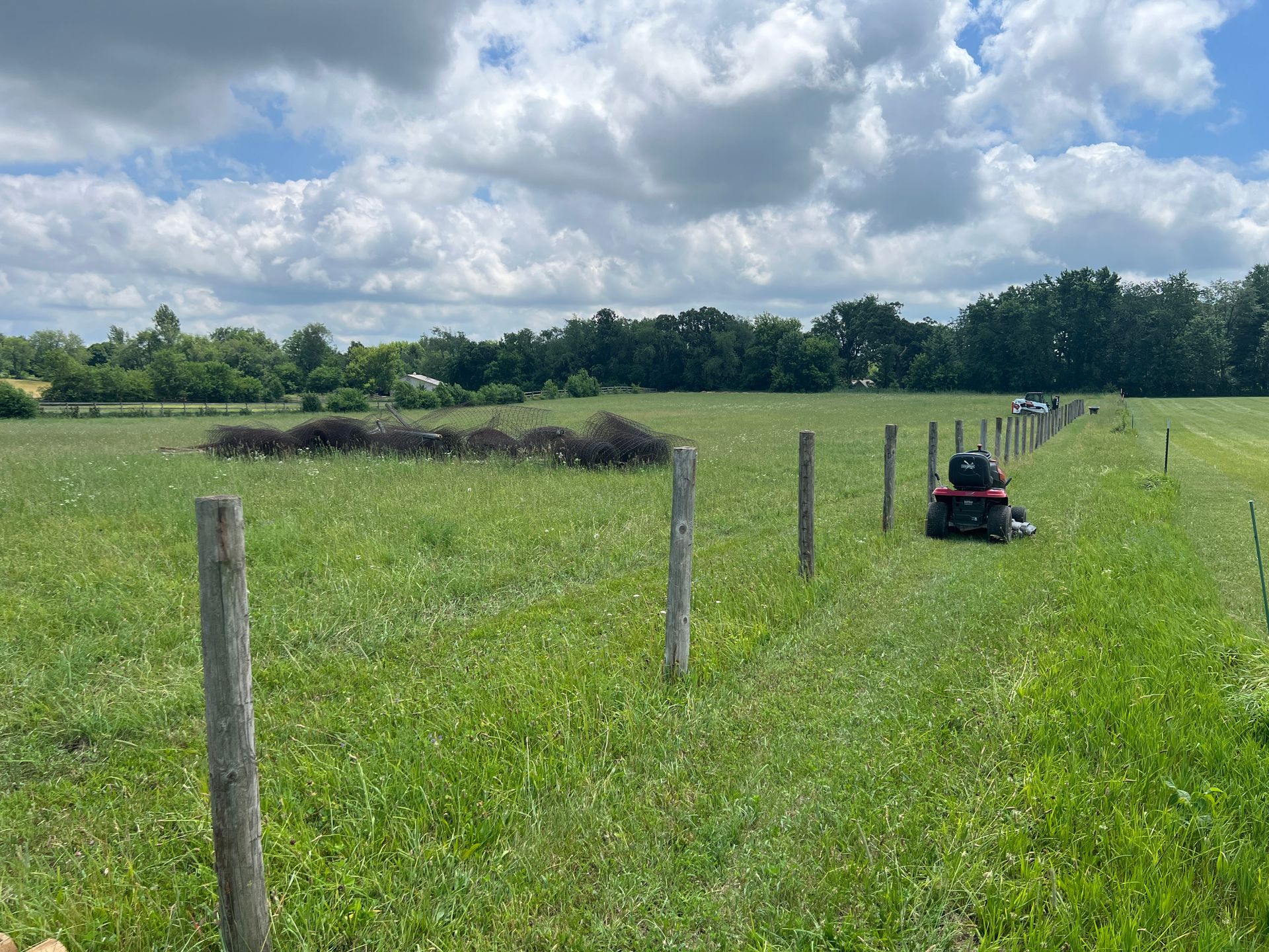 A red riding mower sits along a wooden fence line in a large, grassy field under a cloudy blue sky.