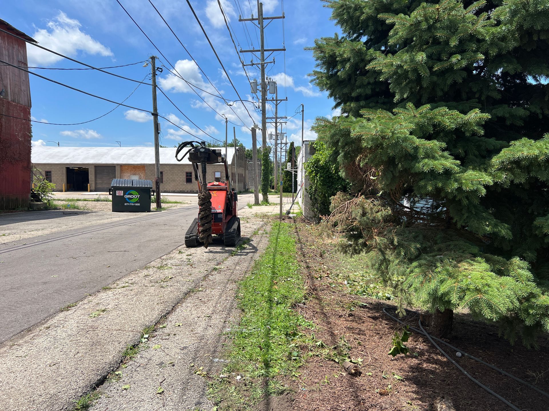 A bright red trenching machine operates on a gravel shoulder next to a paved road, utility poles, and a large conifer.