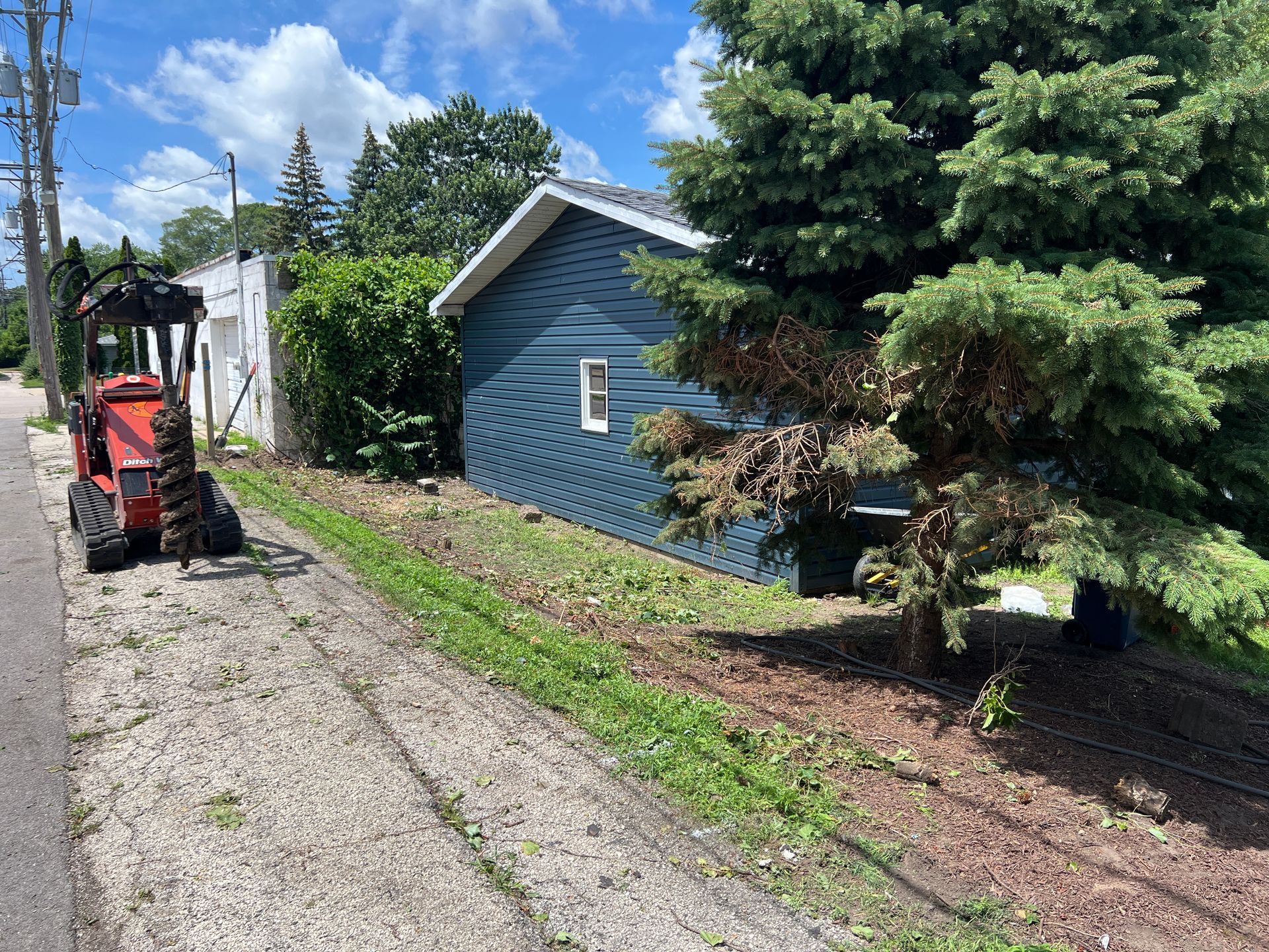 A compact track loader with an auger attachment sits on a gravel path next to a small blue house and a evergreen tree.