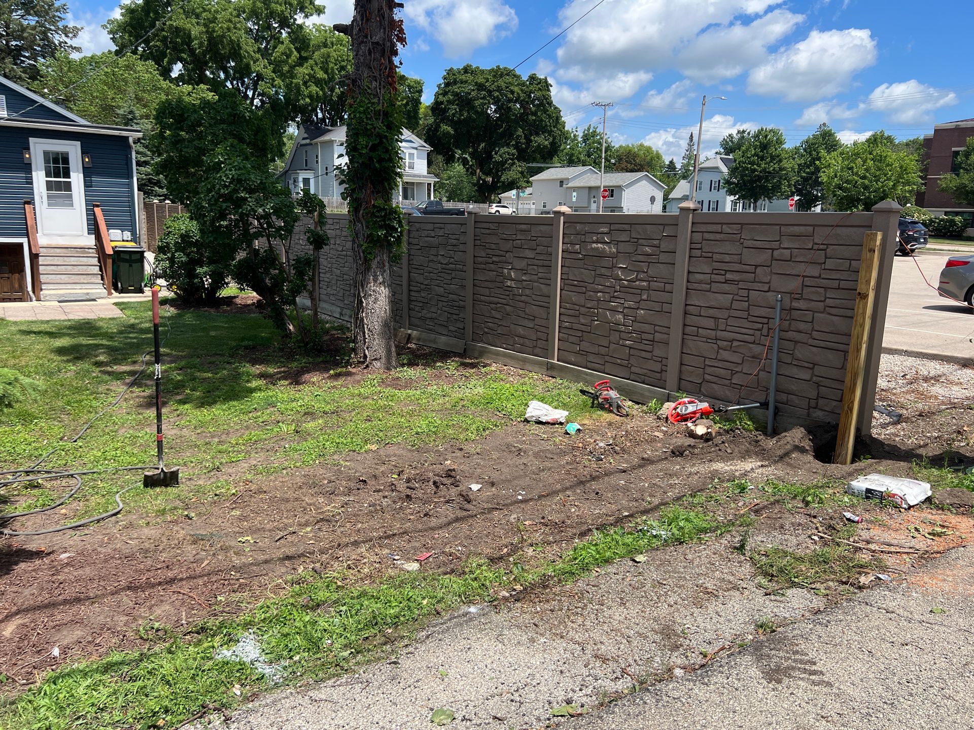 A residential backyard featuring a textured, gray fence, a large tree, and a garden bed under construction on a sunny day.