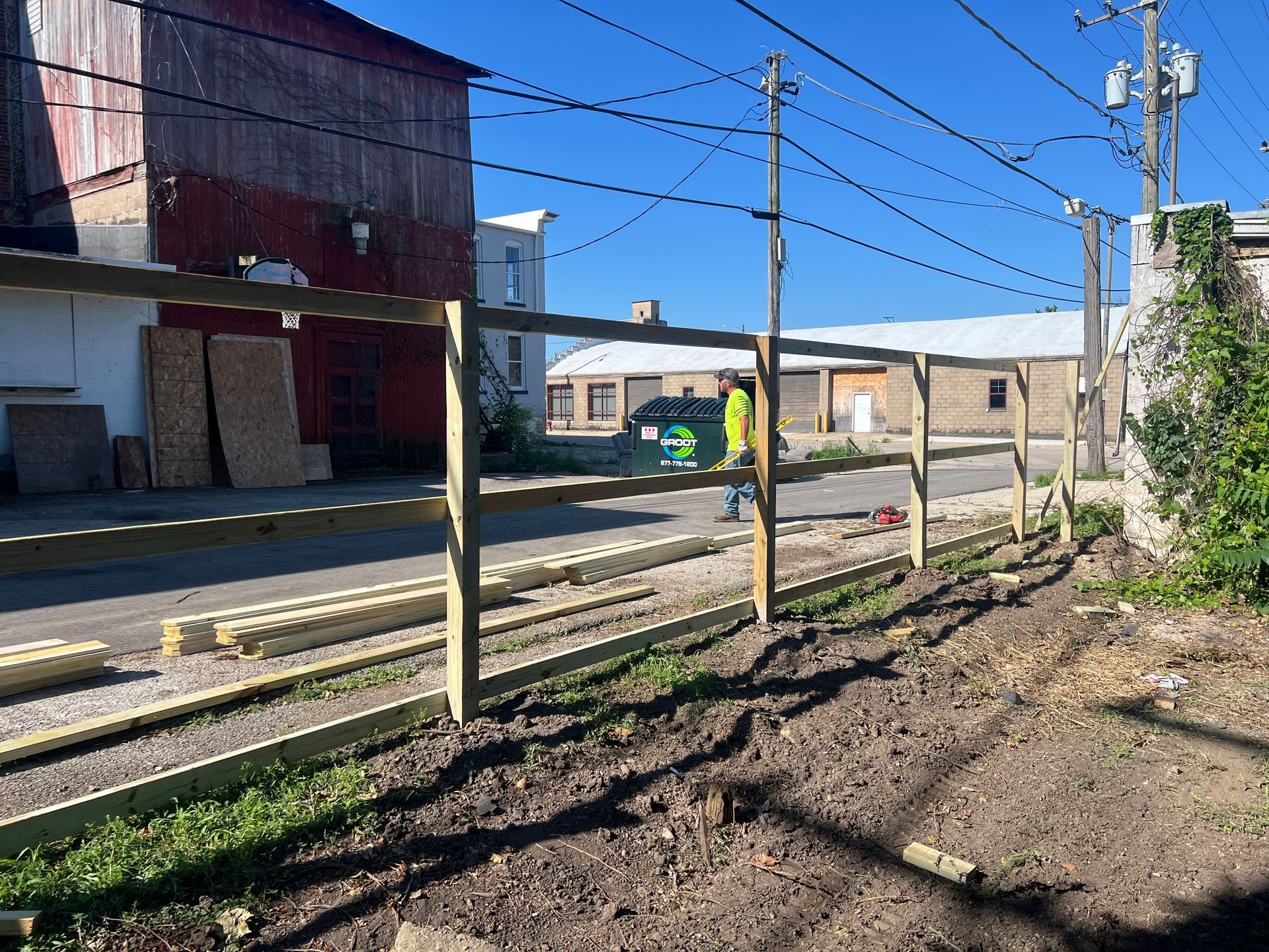 A worker in a high-visibility yellow jacket builds a wooden split-rail fence in a sunny gravel and dirt outdoor lot.