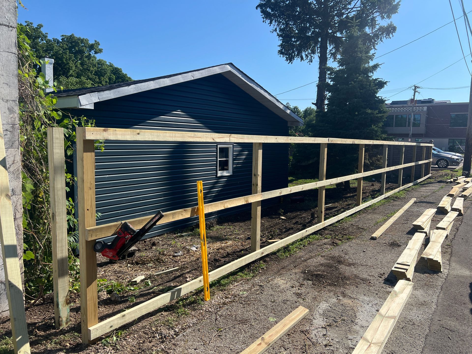 A new wooden fence frame stands in a sunny backyard, positioned in front of a house with dark blue horizontal siding.