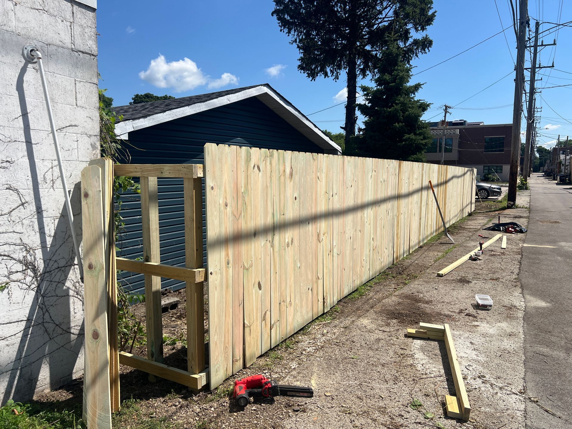 A new wooden fence stands under construction next to a building and an alleyway on a sunny day.