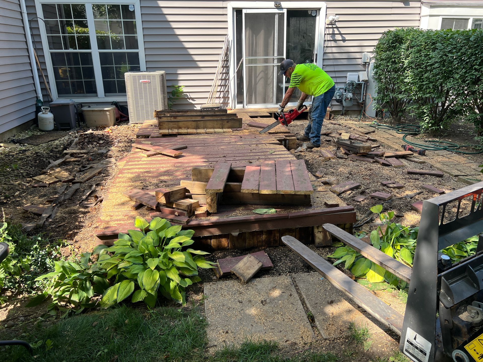 A person in a neon yellow shirt uses a power saw to dismantle a weathered wooden deck in a residential backyard.