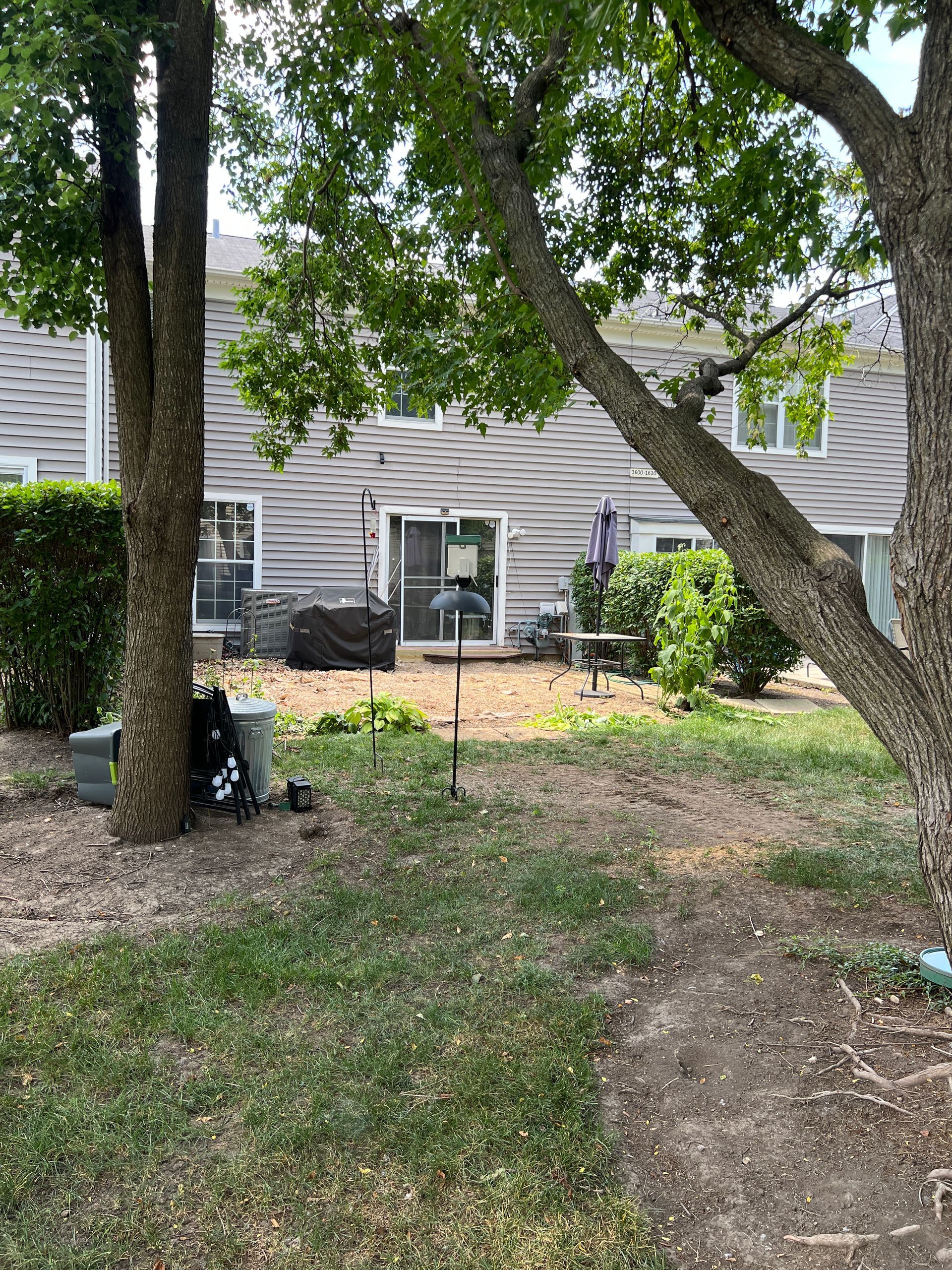 A backyard view showing a patio with a grill and outdoor table behind two large trees, framed by green lawn and foliage.