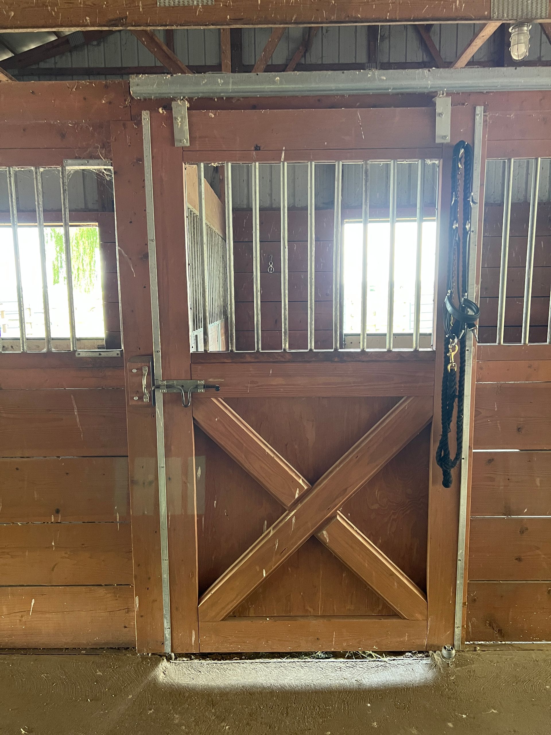 A brown wooden horse stall door with a sliding metal grate and a black bridle hanging on the right side.