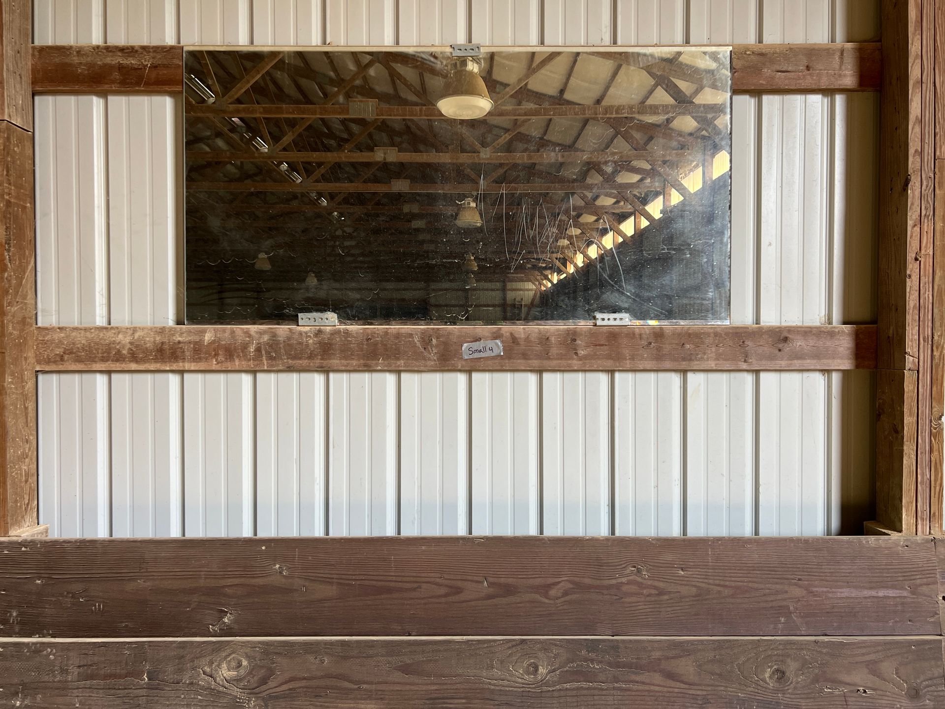 A large mirror mounted on a white corrugated metal wall between wooden barn supports, reflecting the indoor arena ceiling.