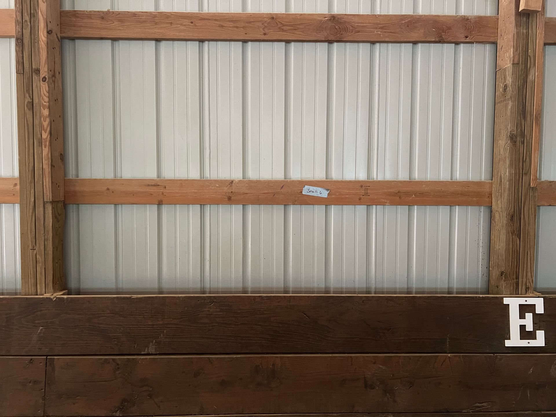 A stall wall with vertical white metal siding, horizontal wooden support beams, and a dark wood lower panel labeled with 