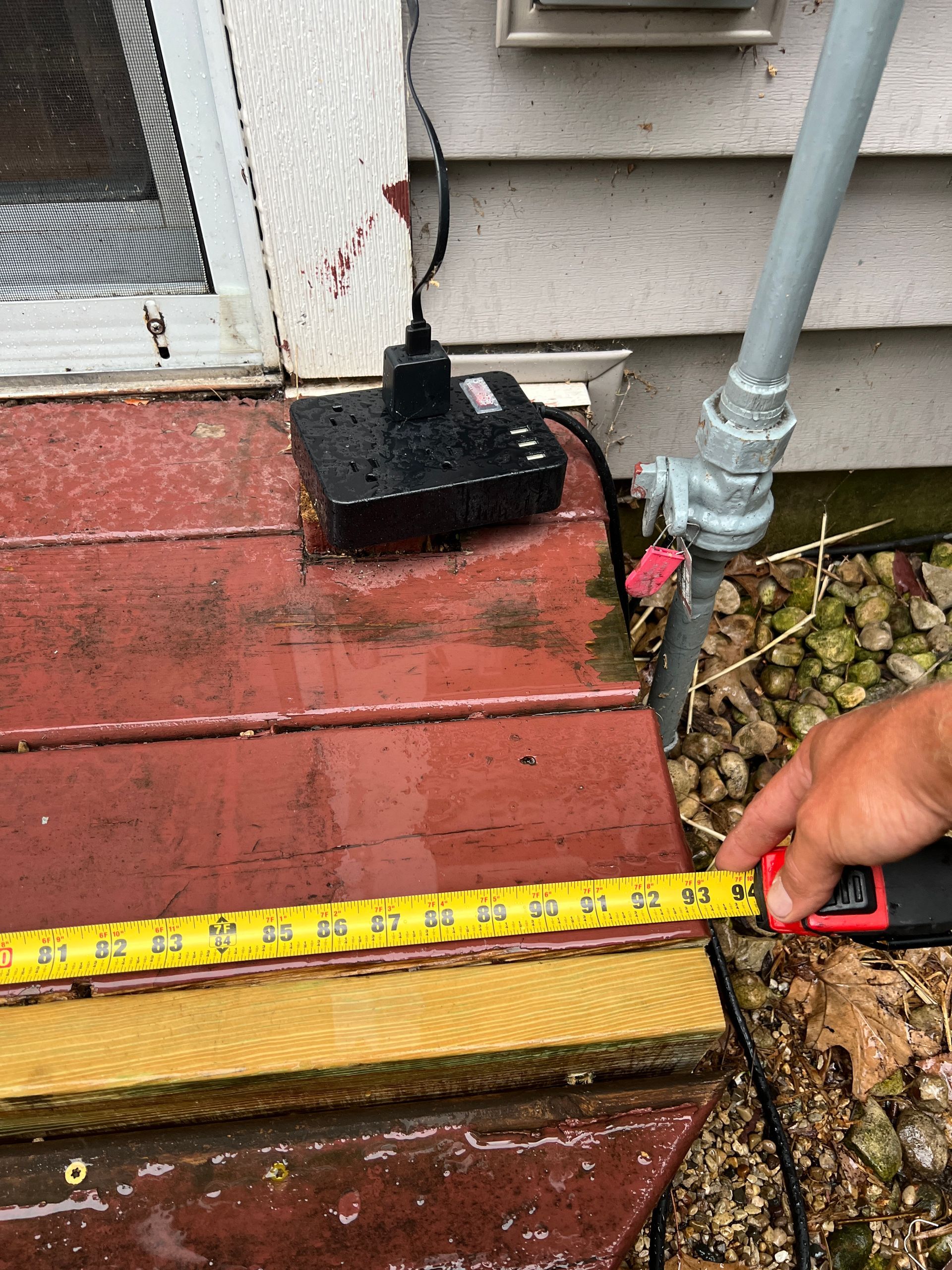 A person uses a yellow tape measure to measure the width of a red wooden deck next to an outdoor electrical power strip.