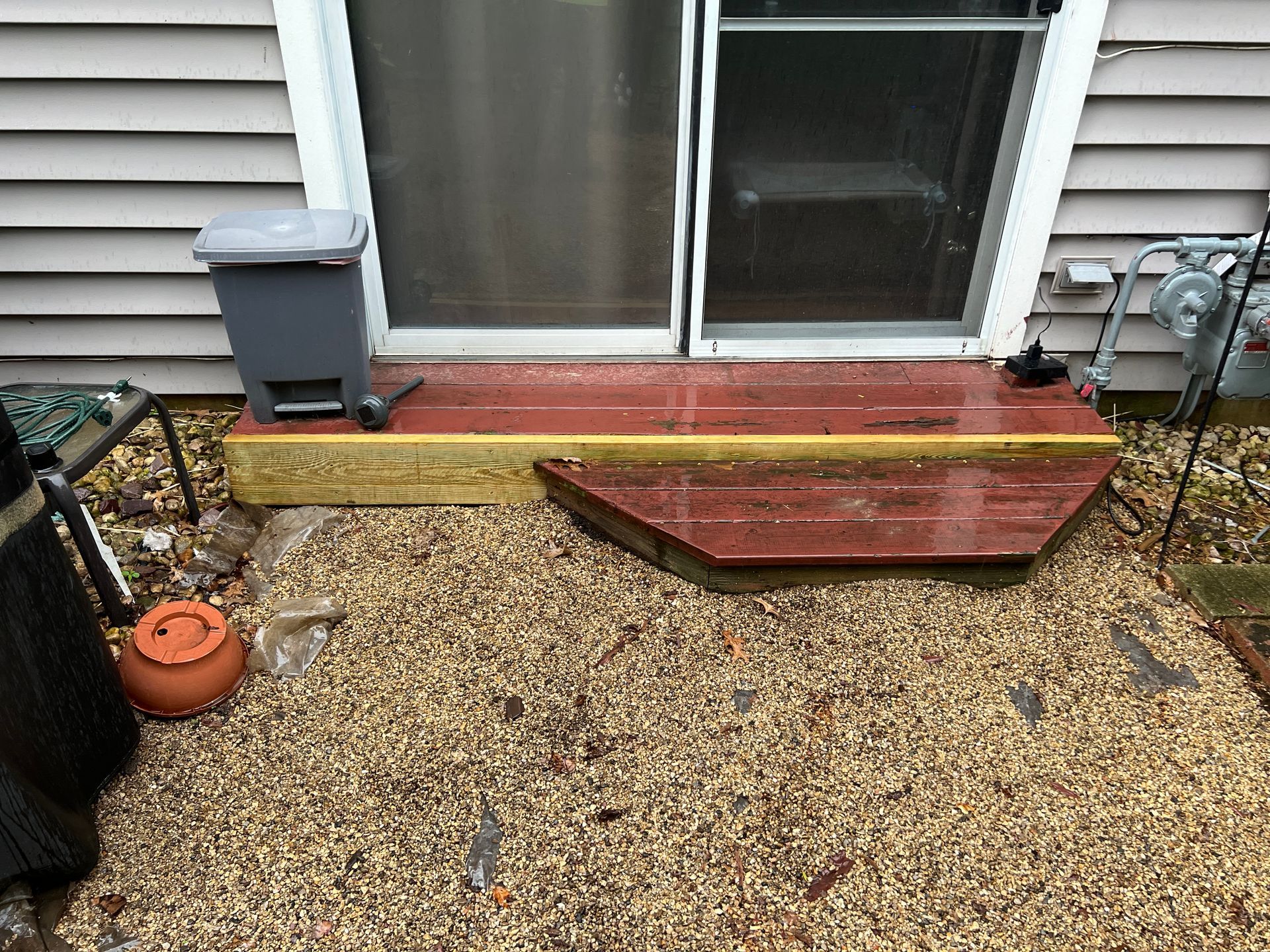 A small wooden step and landing outside a sliding glass door with a gravel ground area and a small grey trash bin.