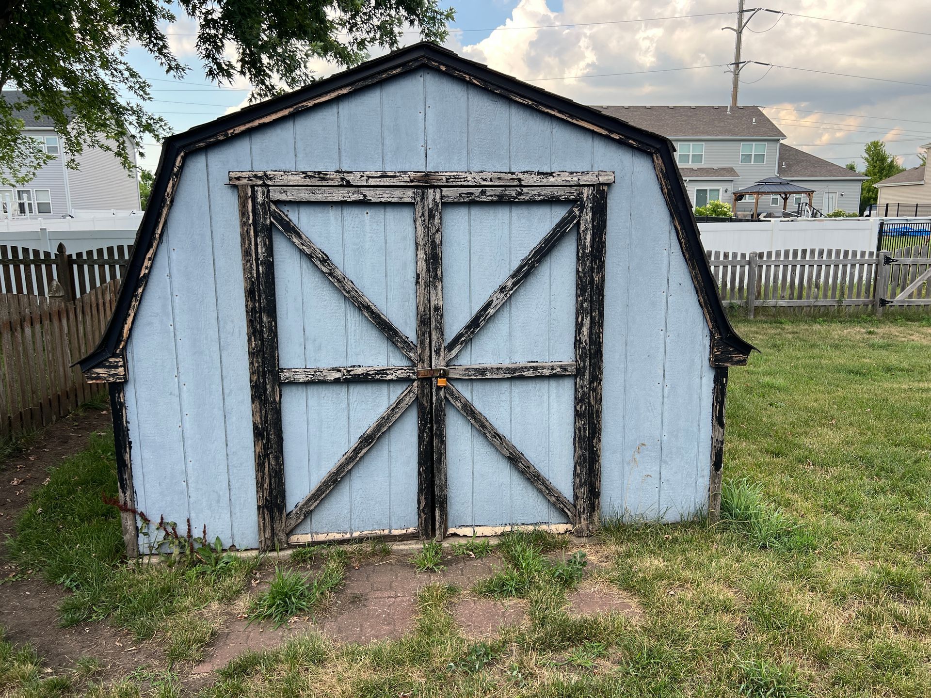 A light blue wooden shed with black trim and double doors featuring an X-pattern sits in a grassy backyard.