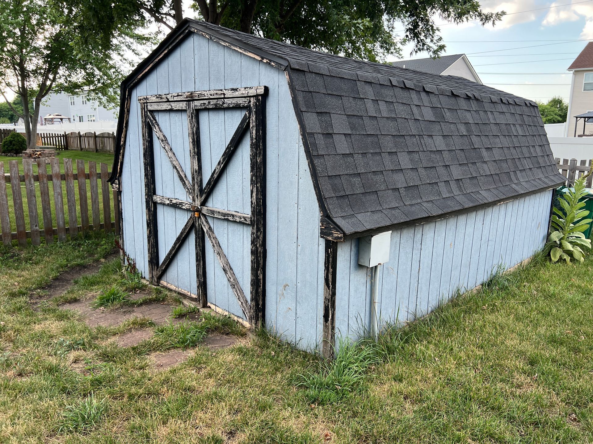 A weathered, light blue wooden storage shed with a gambrel roof and dark trim, situated on a grassy lawn.