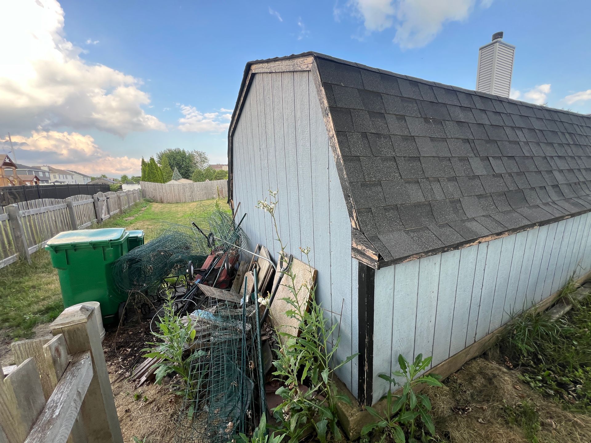 A light blue shed in a backyard with a pile of scrap, a green bin, and a fence under a sunny sky.