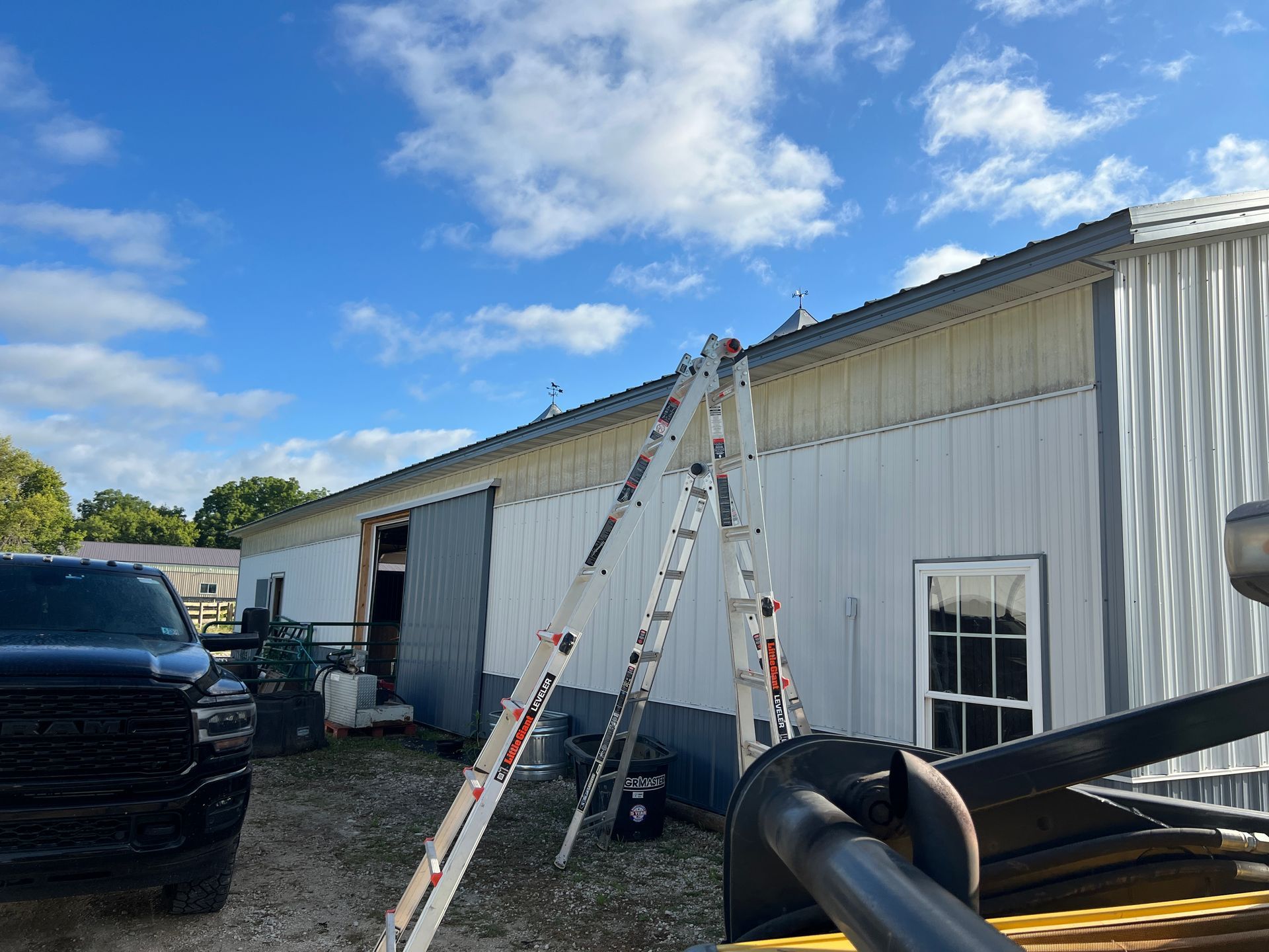 A tall extension ladder leans against the exterior wall of a white metal building under a blue, partly cloudy sky.