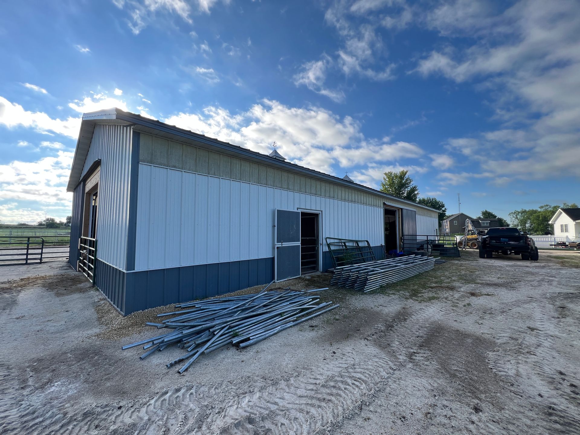 A long, white and dark gray pole barn on a dirt lot with metal fencing stacked on the ground under a blue, cloudy sky.