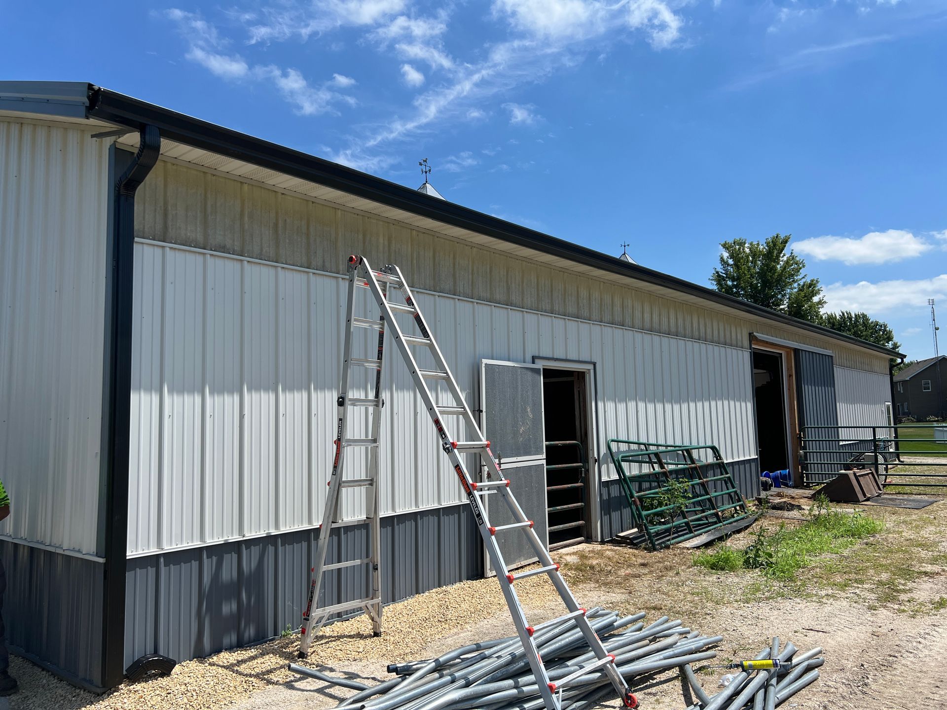 A long metal building with light-colored siding, a dark grey base, and a ladder leaning against the wall outside.