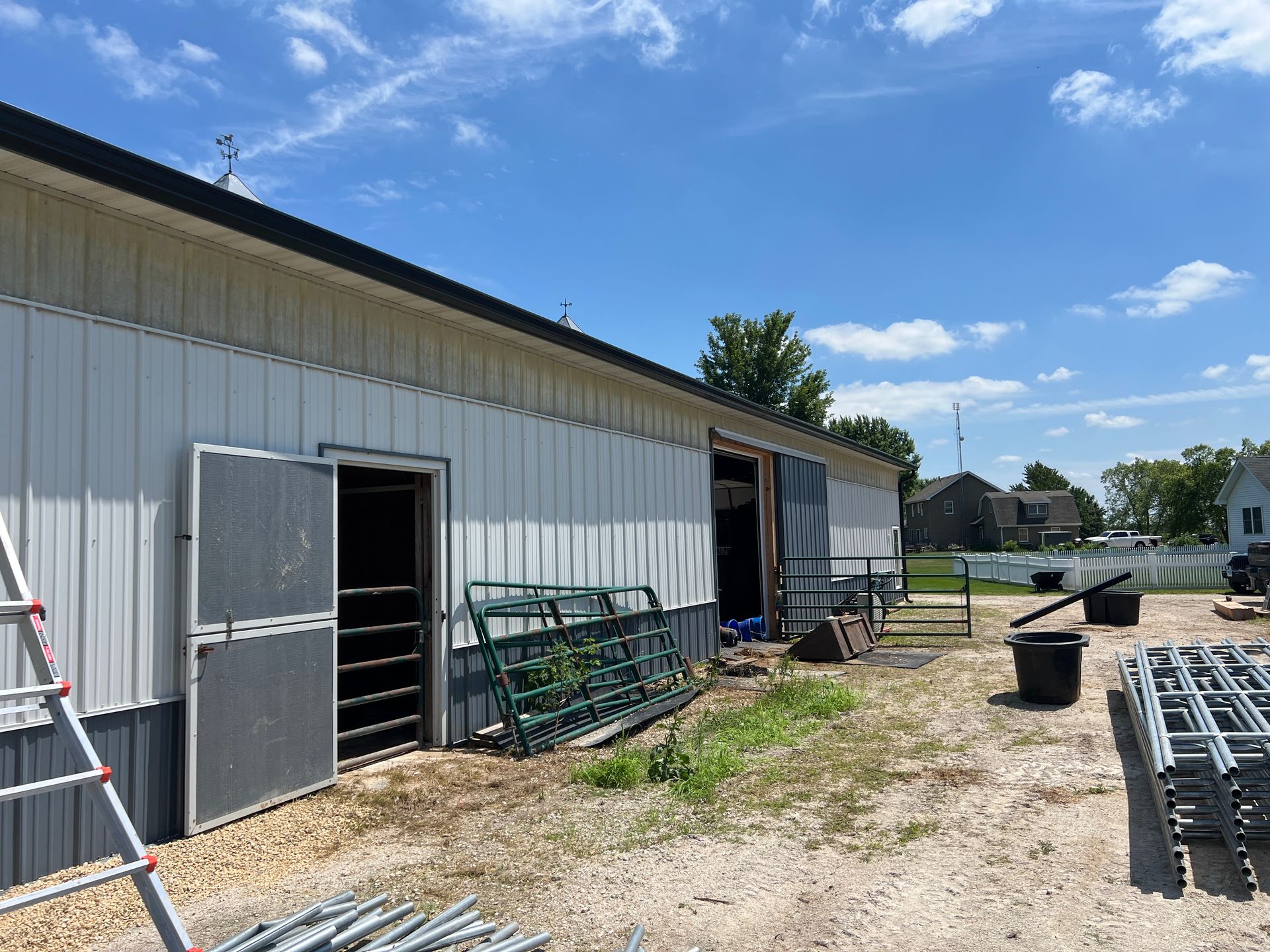 Exterior of a pole barn with metal siding, horse stall doors, and various metal farm equipment panels on a gravel lot.