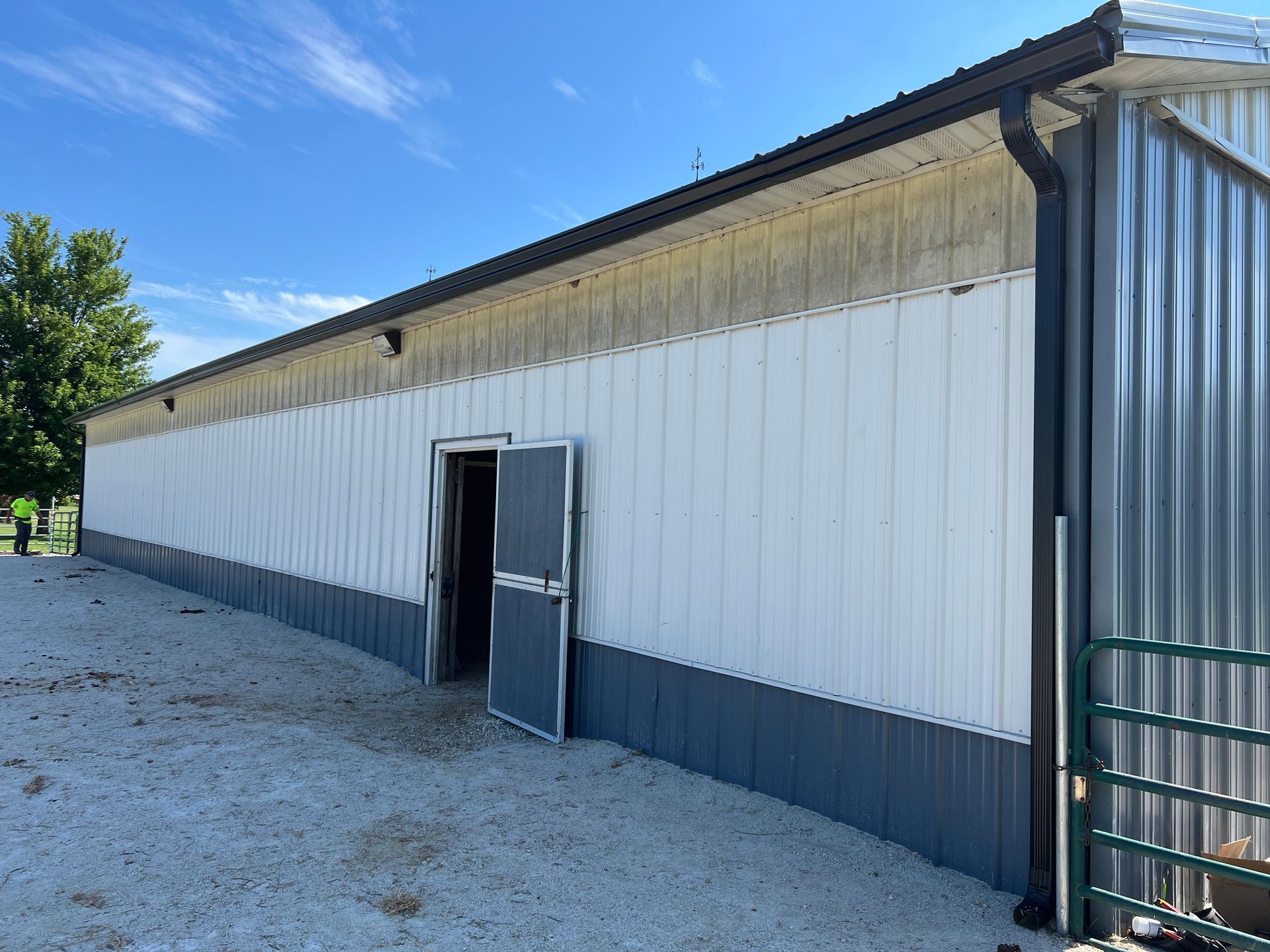 A long metal barn with white walls, dark gray trim, and an open doorway on a gravel lot under a blue sky.