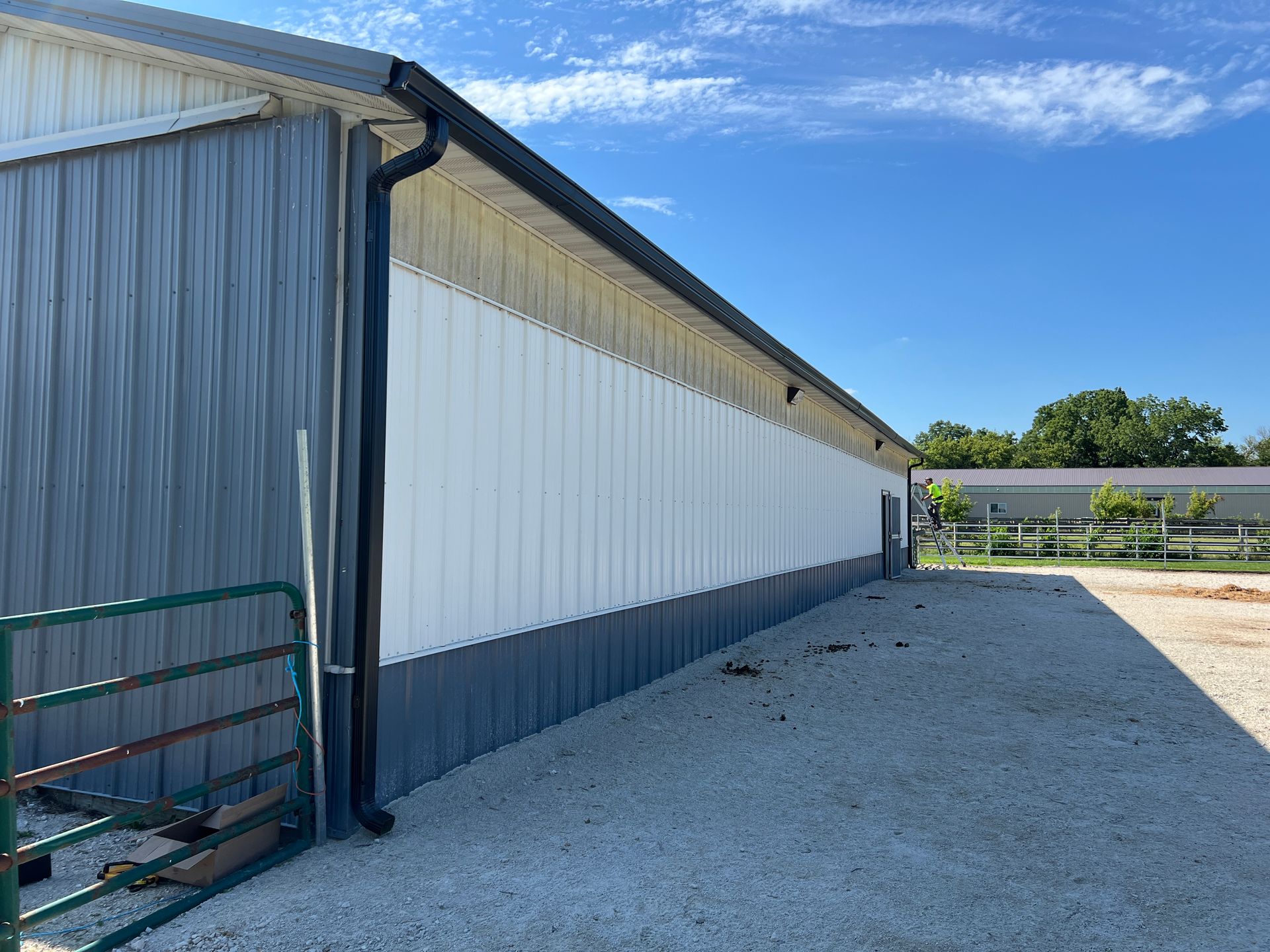 A long, metal-sided agricultural building with grey lower walls, white upper panels, and a black downspout on gravel.