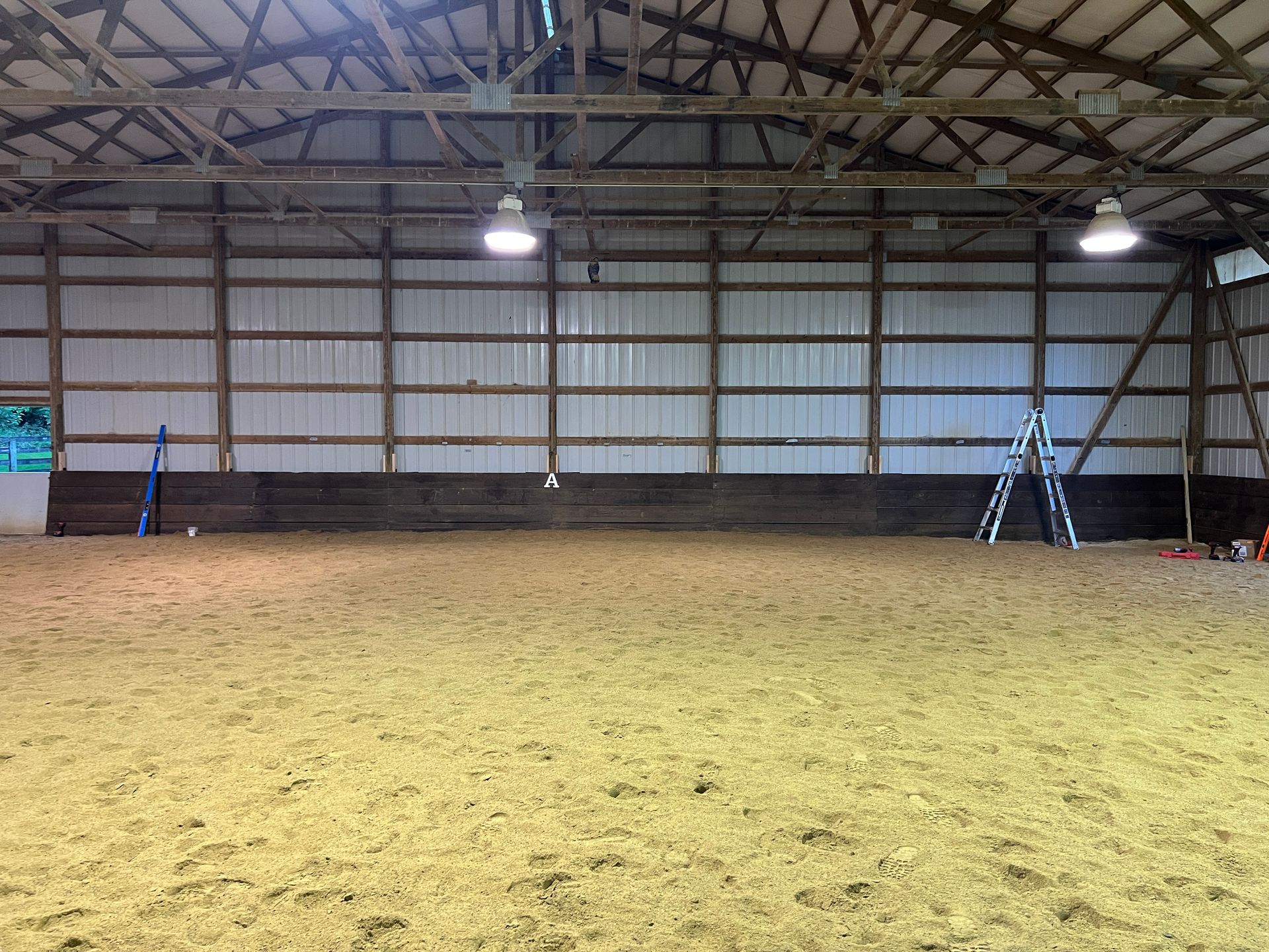 Indoor horse riding arena with a sandy floor, white metal walls, dark kickboards, and a ladder leaning against the wall.