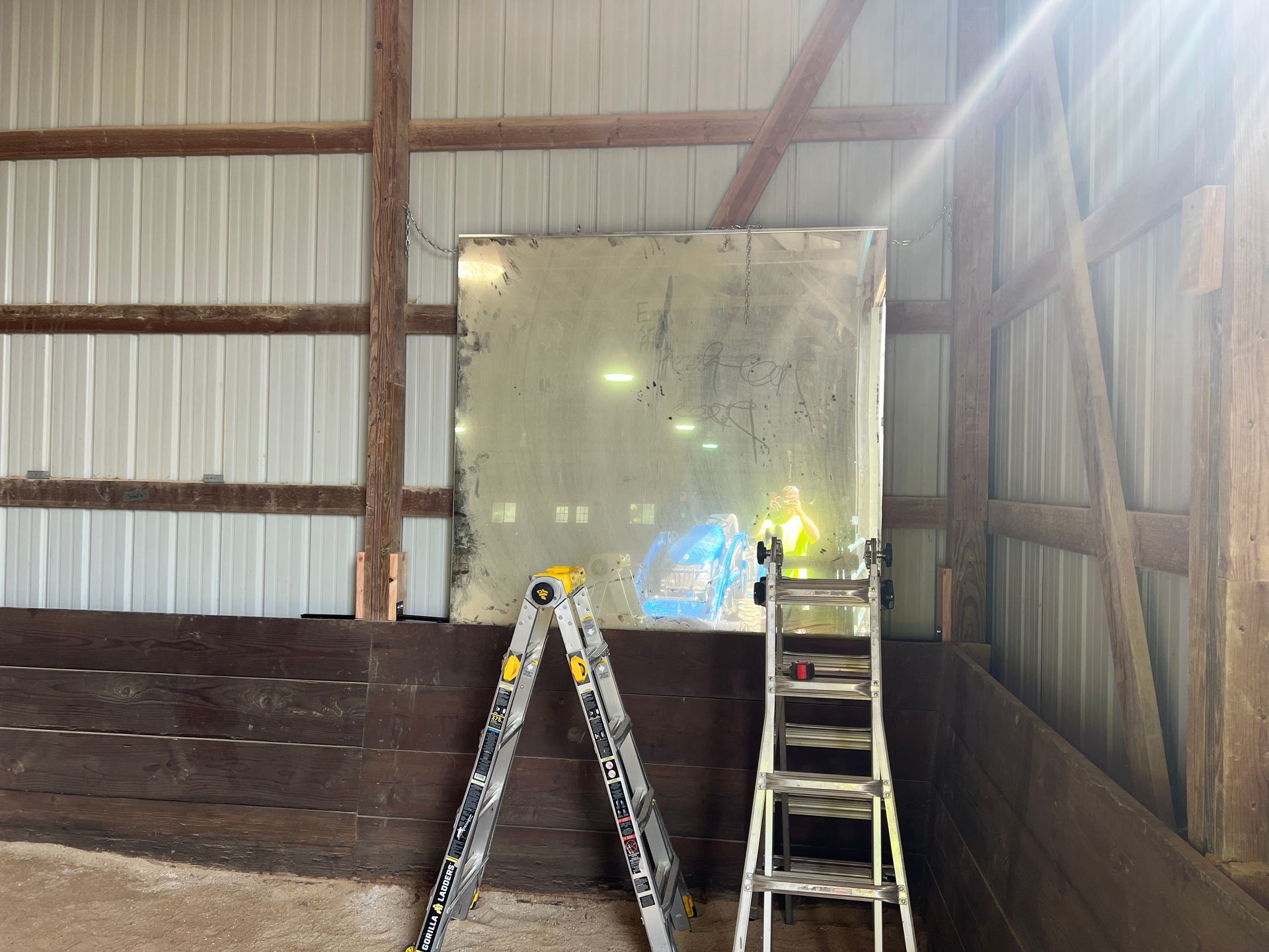 A large, rectangular mirror leans against the white corrugated wall of a rustic indoor arena, next to two ladders.