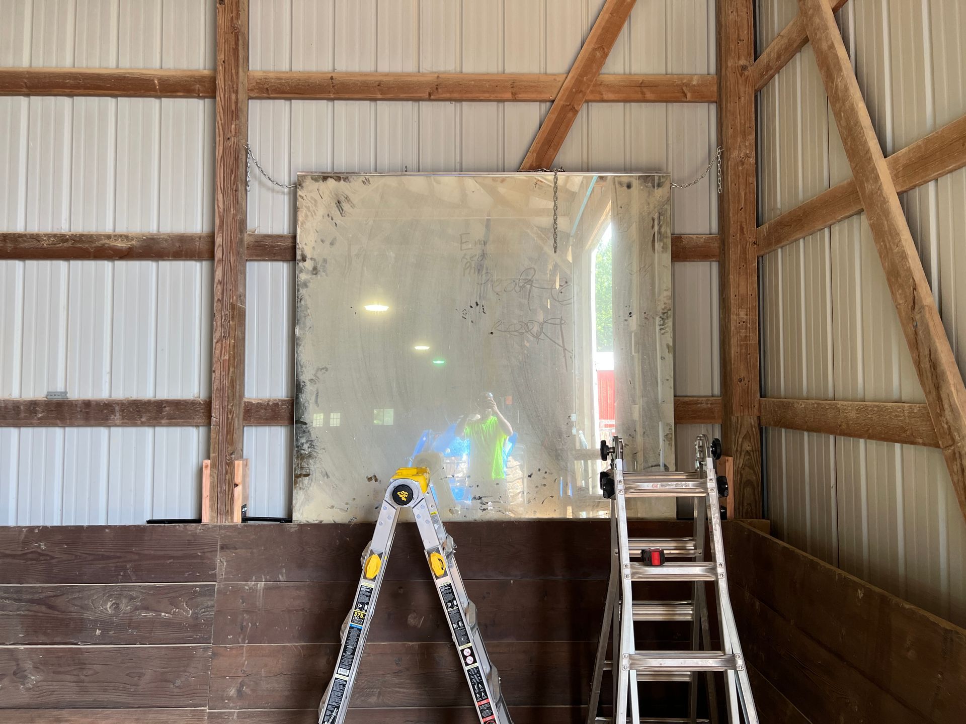 A large, weathered rectangular mirror leaning against a wood-paneled wall next to a folding ladder in a barn.