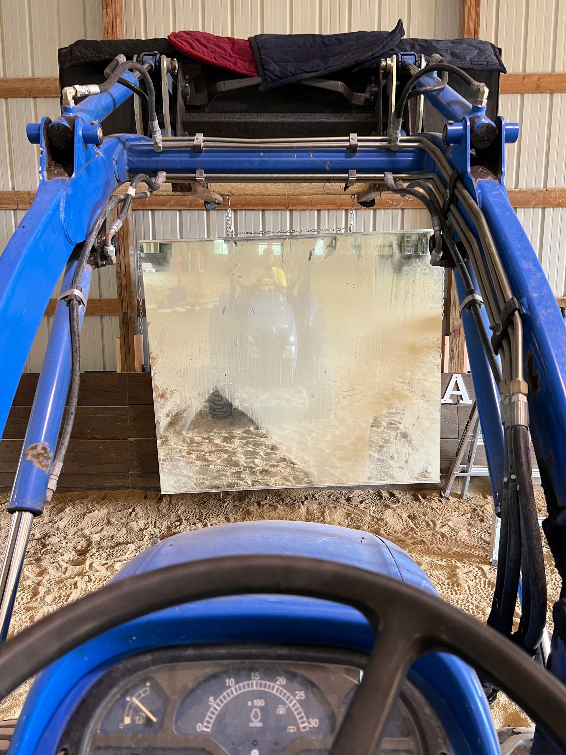 A view from the driver's seat of a blue tractor, showing its loader arms framing a large, dirty mirror in a barn.