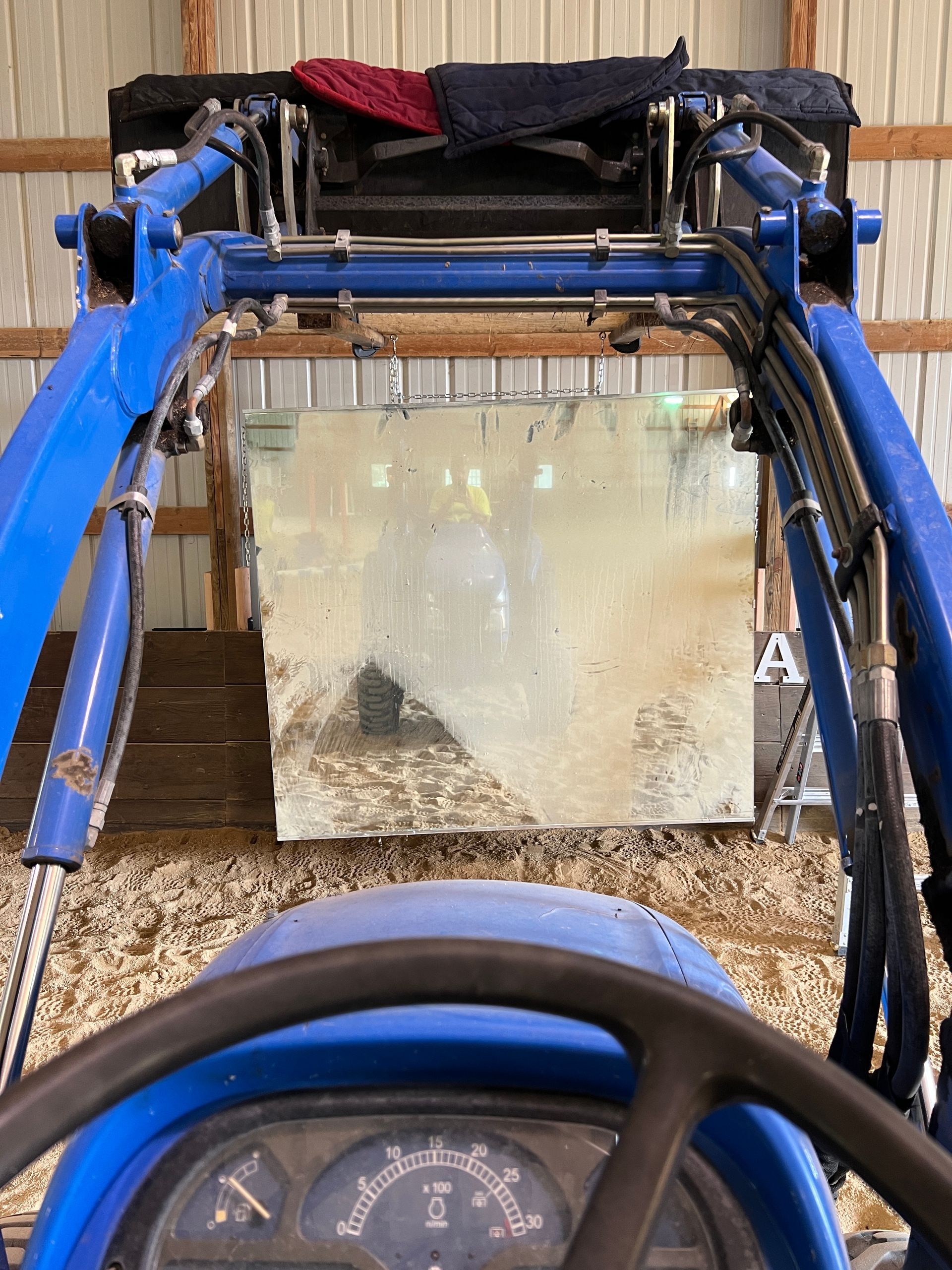 View from the driver's seat of a blue tractor, facing a large, square, light-colored pane of glass in an indoor barn.