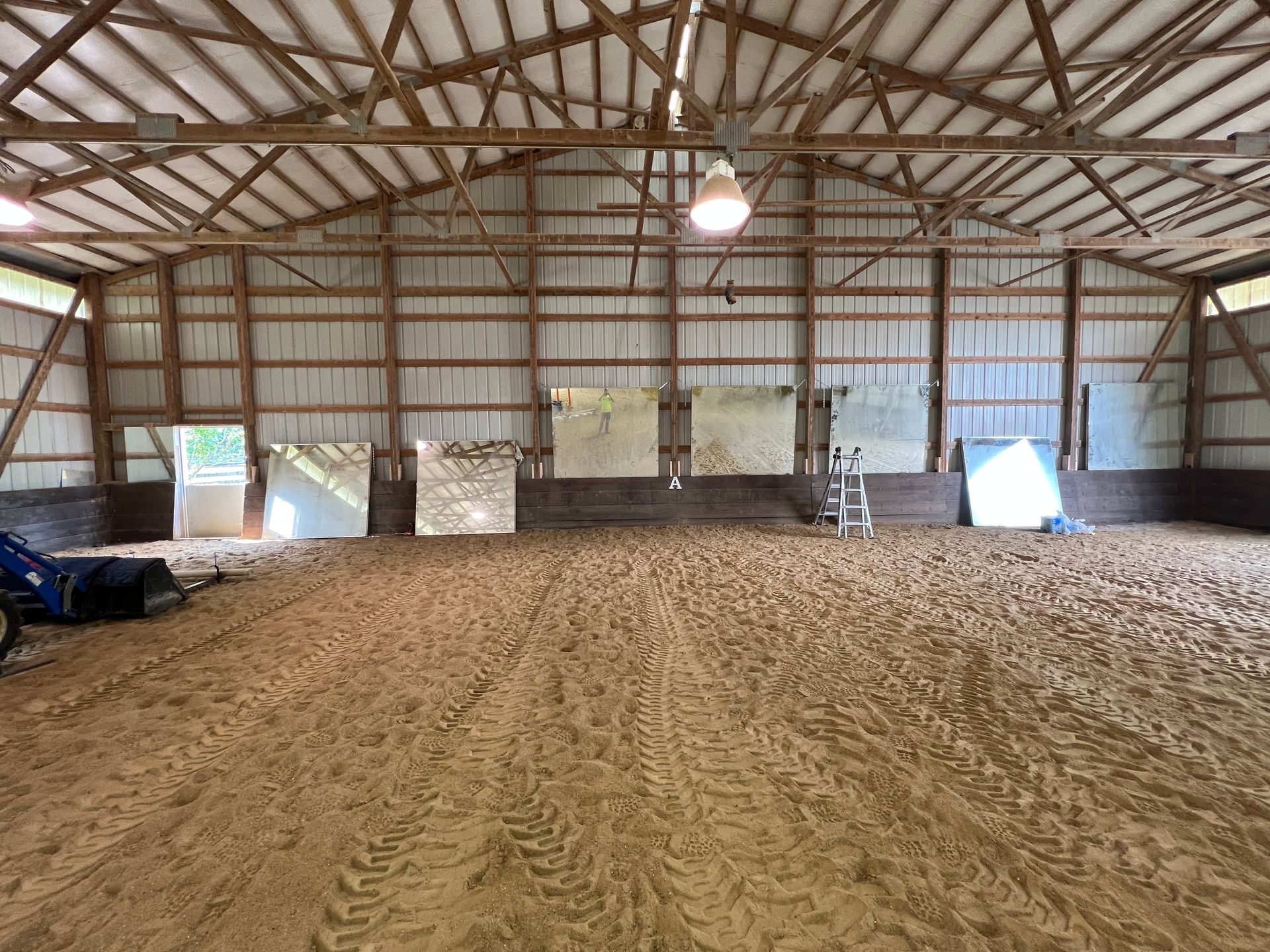 A large, indoor equestrian arena with a sandy floor, wooden walls, exposed ceiling trusses, and hanging mirrors.