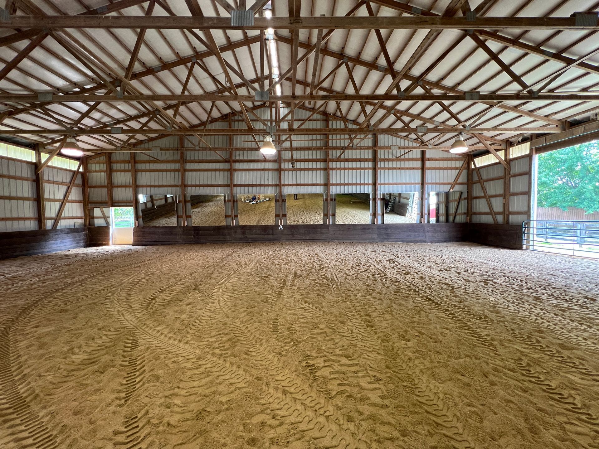 An indoor equestrian riding arena with sand footing, a wooden perimeter wall, and a high, exposed truss roof.