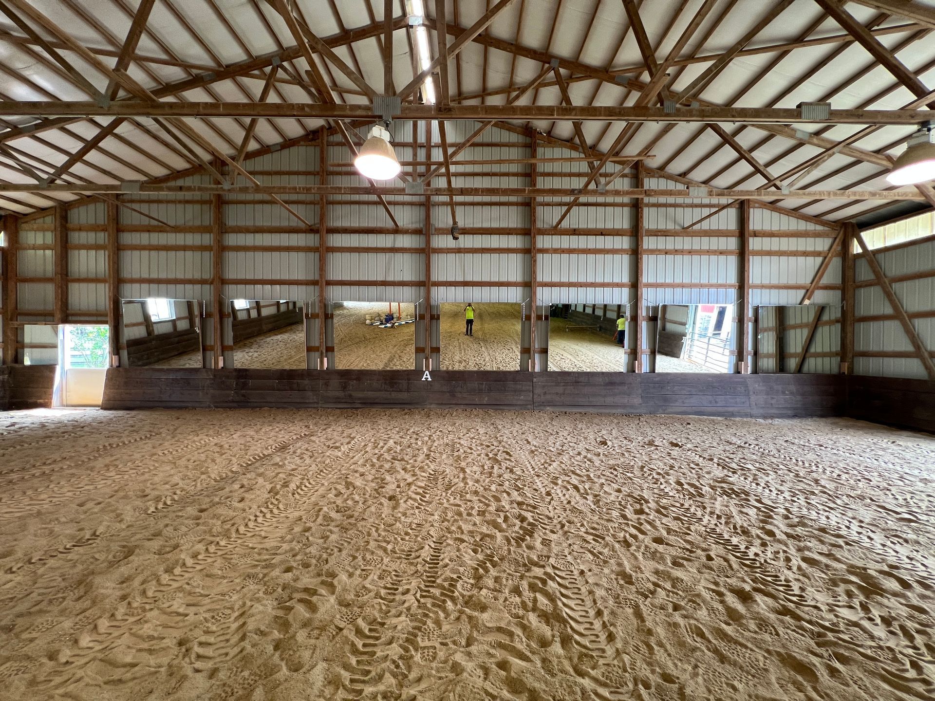 Indoor horse riding arena with sandy ground, wooden kickboards, and mirrors lining the back wall under a high-beam ceiling.