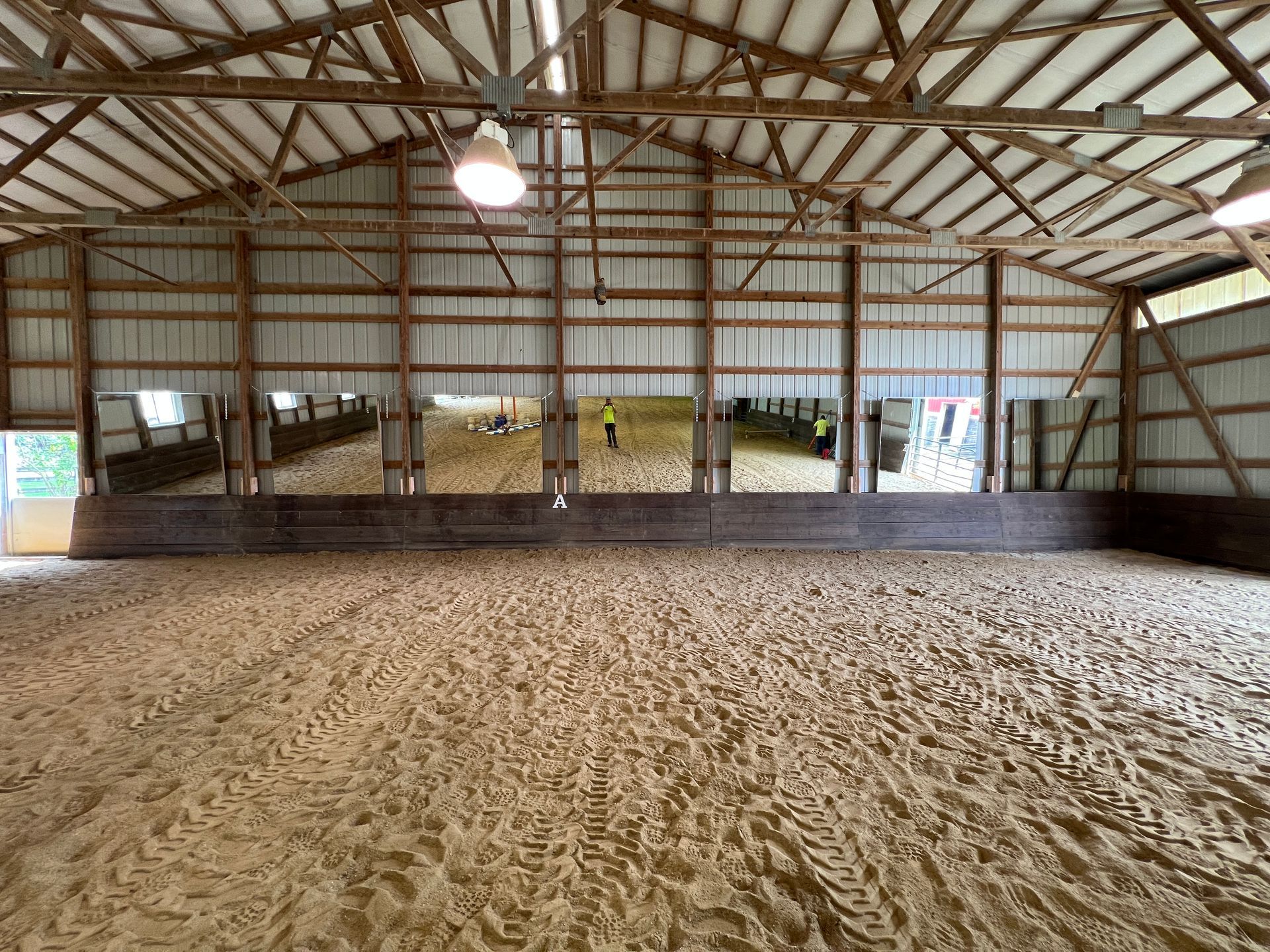 An indoor riding arena with a sandy surface, wooden kickboards, and mirrors reflecting a person in a yellow shirt.