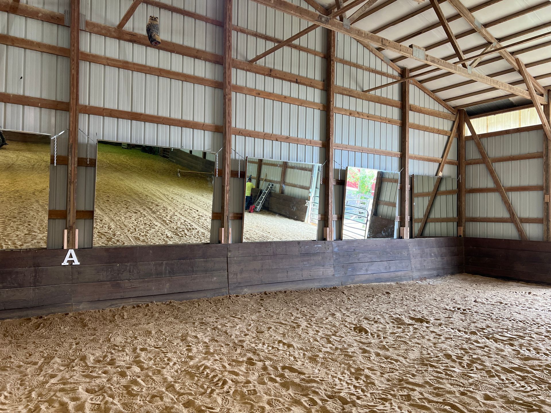 Indoor riding arena with large mirrors mounted on the wall above dark wooden kickboards, reflecting the sandy arena floor.