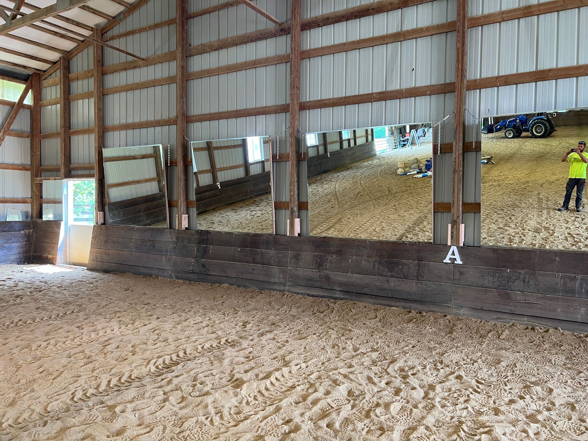 An indoor horse riding arena with sand flooring, wooden kickboards, a row of wall mirrors, and a person in a safety vest.