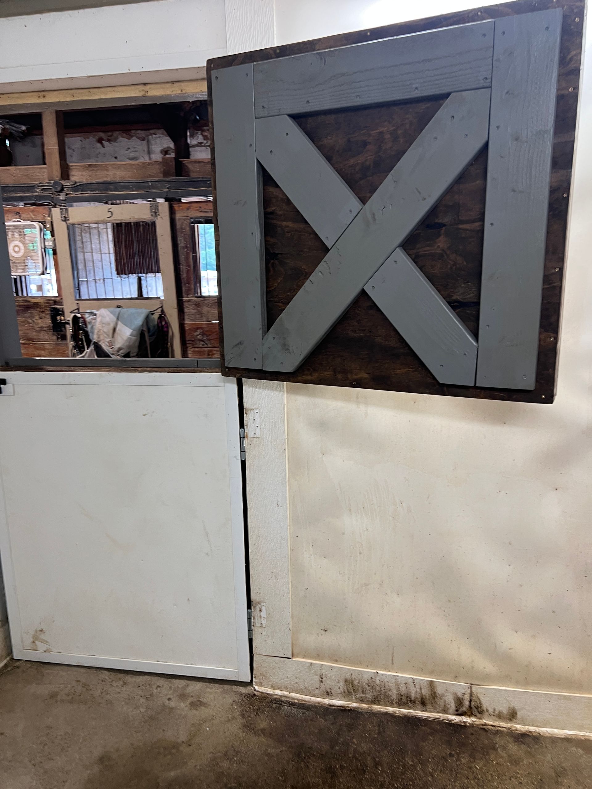 A stable stall with a white bottom door and an open top section featuring a grey X-pattern gate on a dark wooden frame.