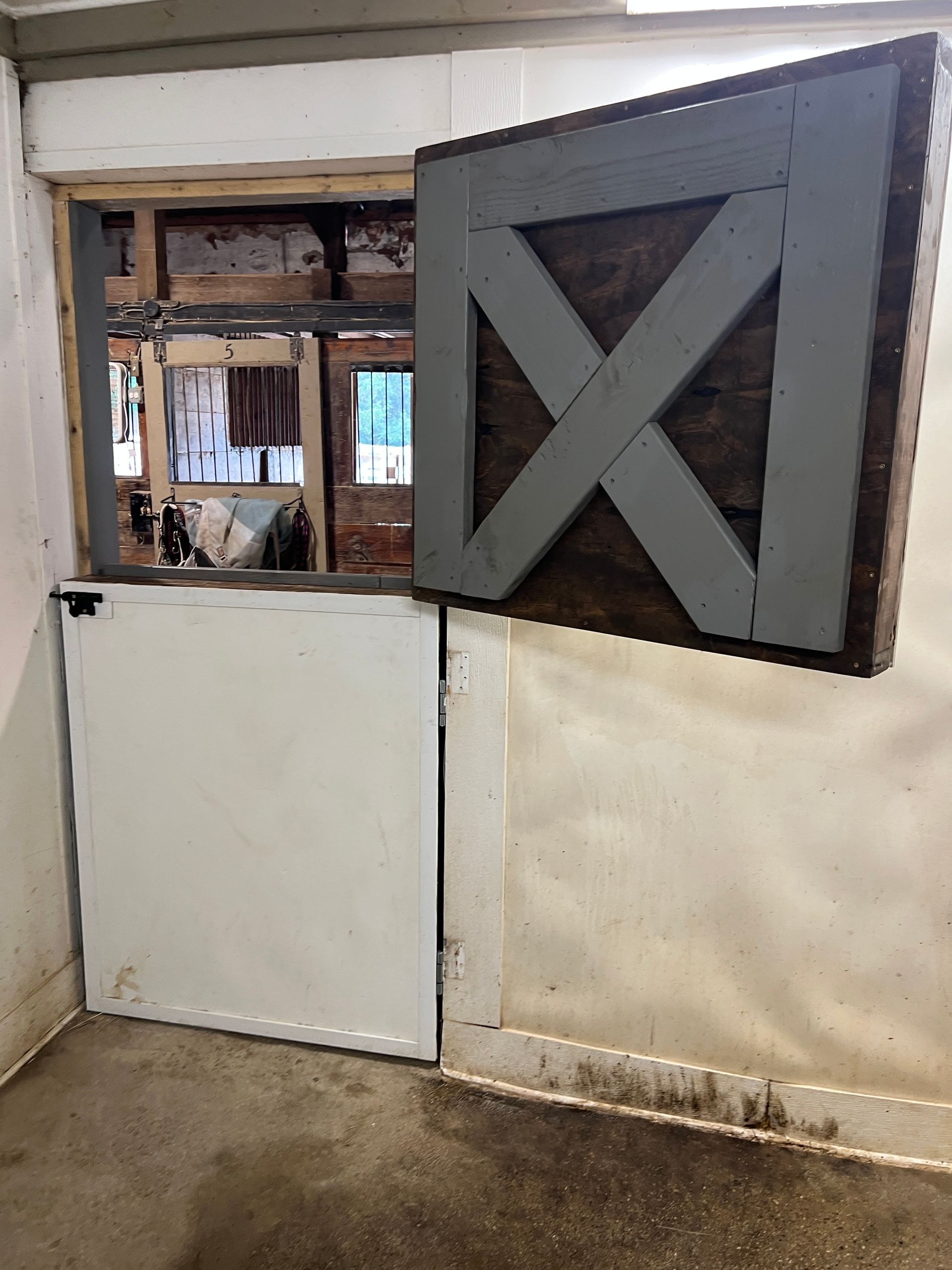 A split-style stable door in a stall: the bottom half is solid white, and the top half is an open grey frame with an X.