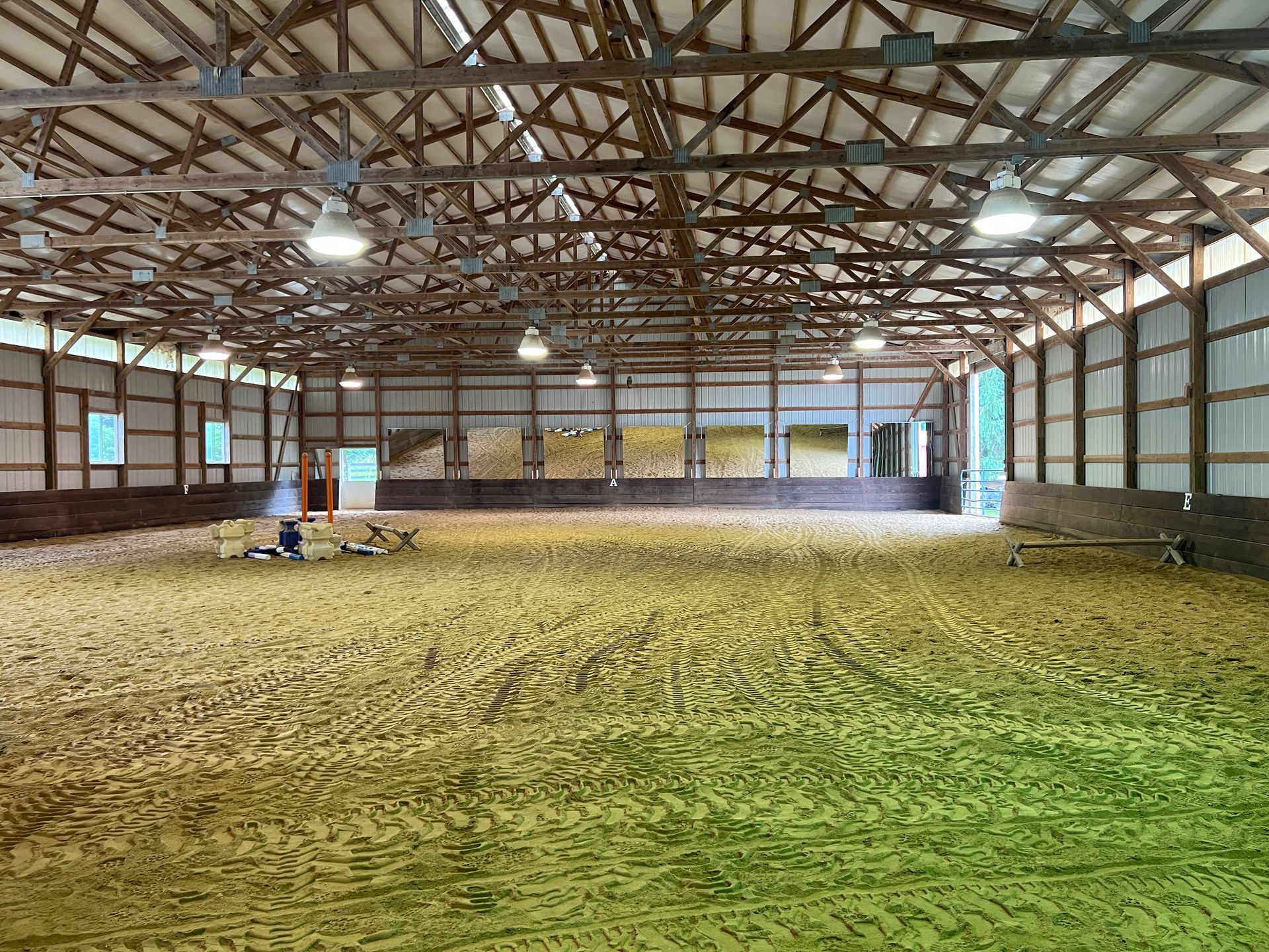 Interior of a large indoor equestrian riding arena with wooden rafters, sand footing, and overhead lighting.
