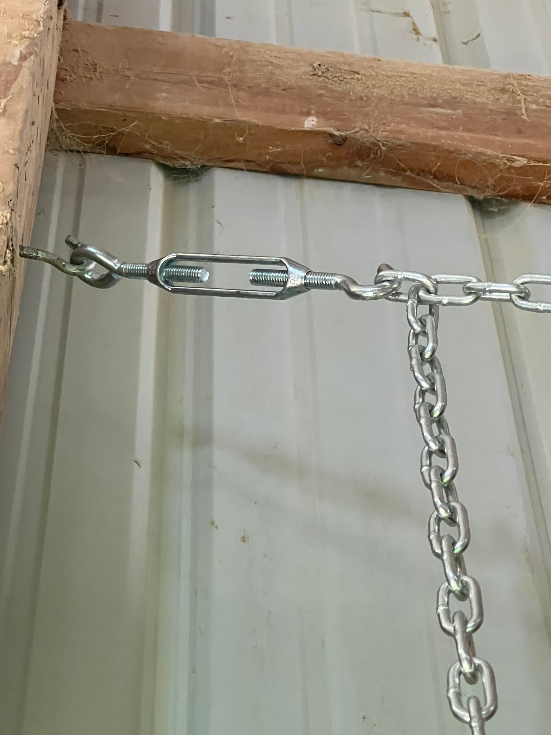 A metal turnbuckle connects a chain to a wooden beam against a corrugated metal wall.