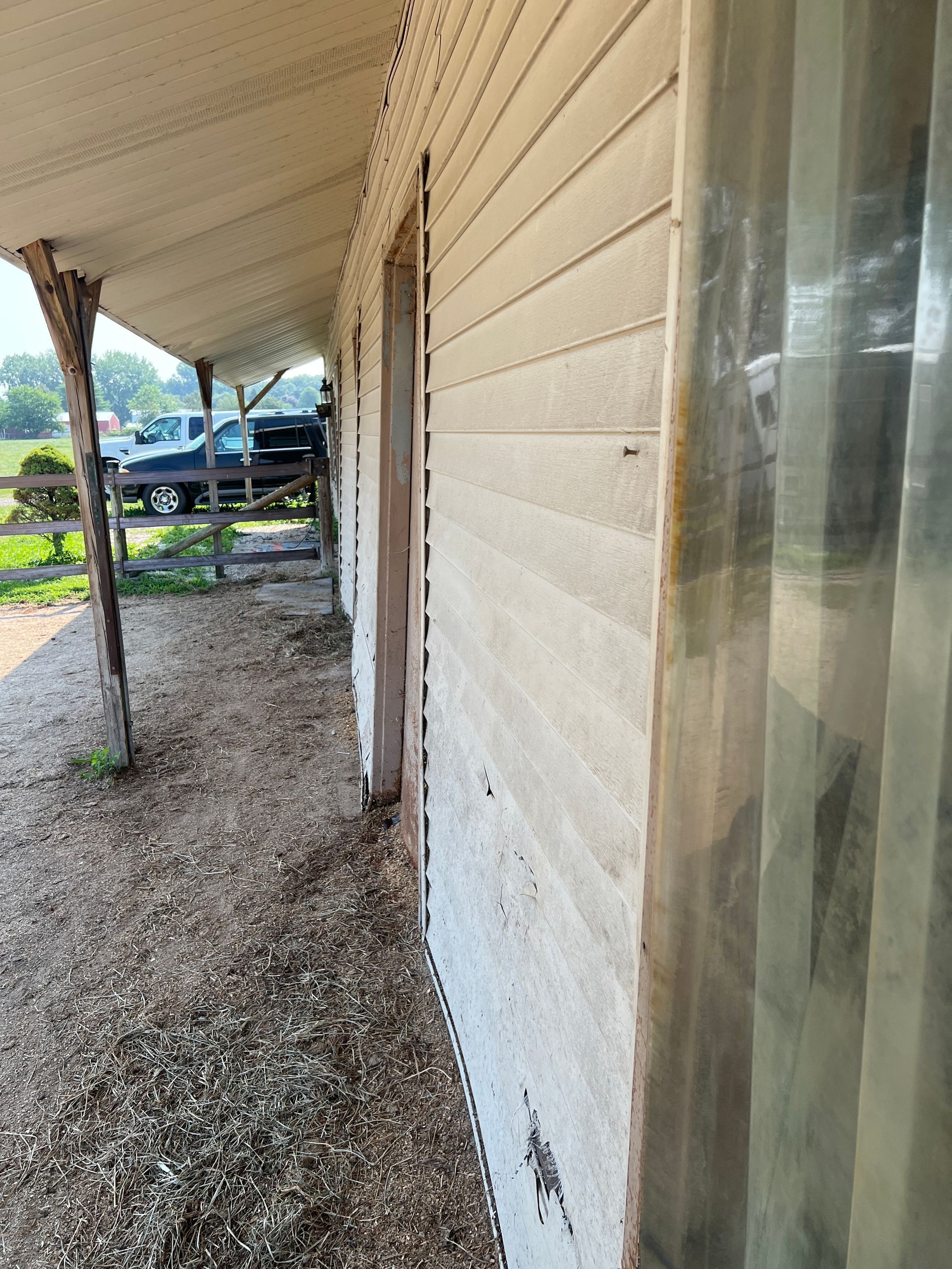 Side view of a porch with beige siding, a wooden support post, and a view of a rural yard with parked vehicles.