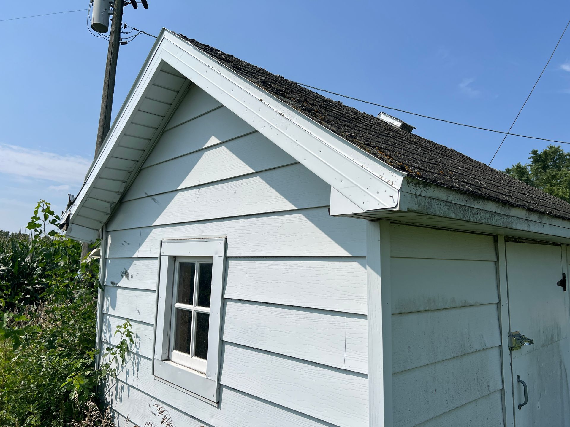 A small, white, wood-sided shed with a gable roof, a single window, and a door under a bright blue sky.