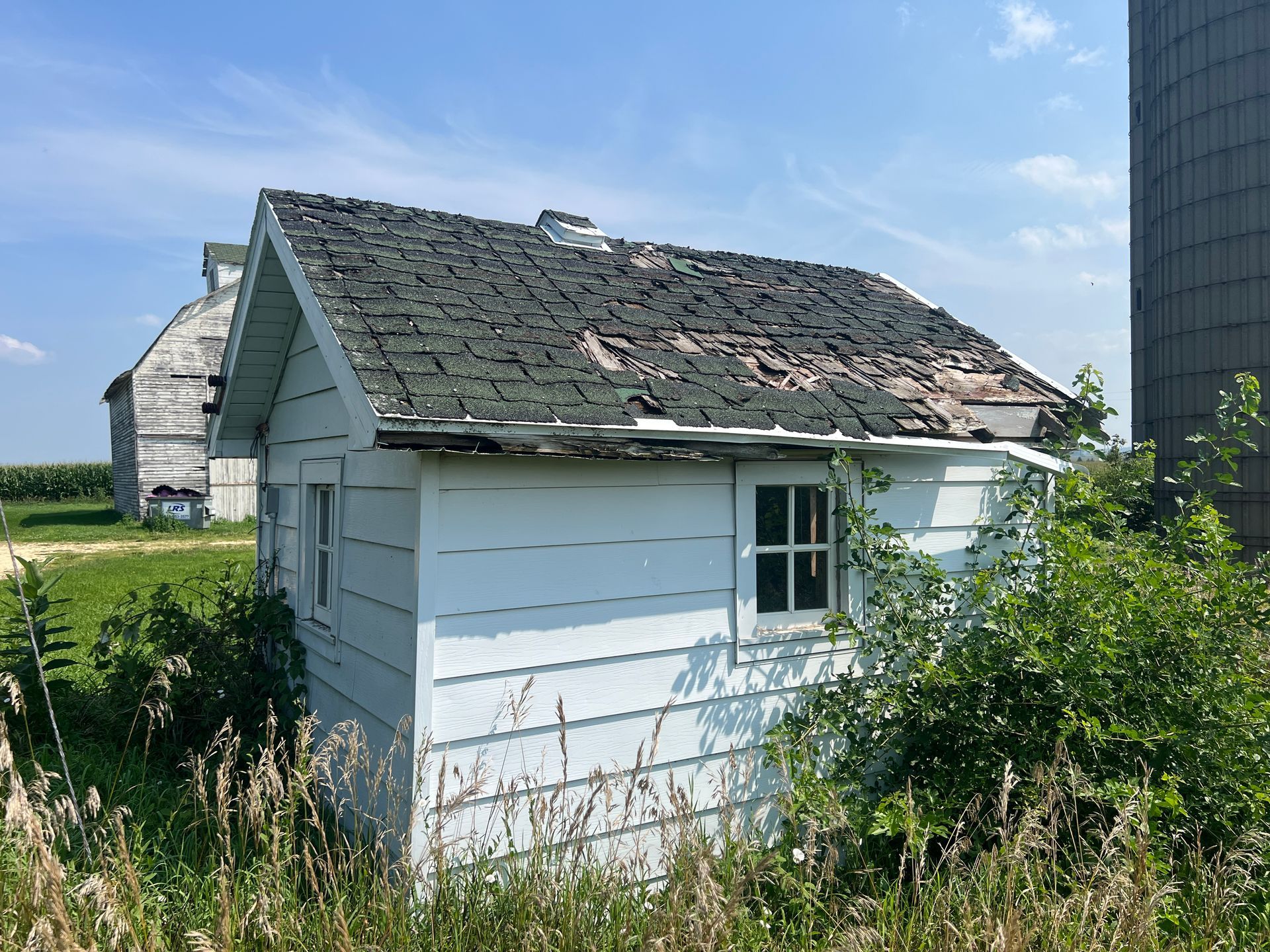 A small, white, weathered shed with a damaged shingle roof sits in a grassy, sunlit field near a farm silo and barn.