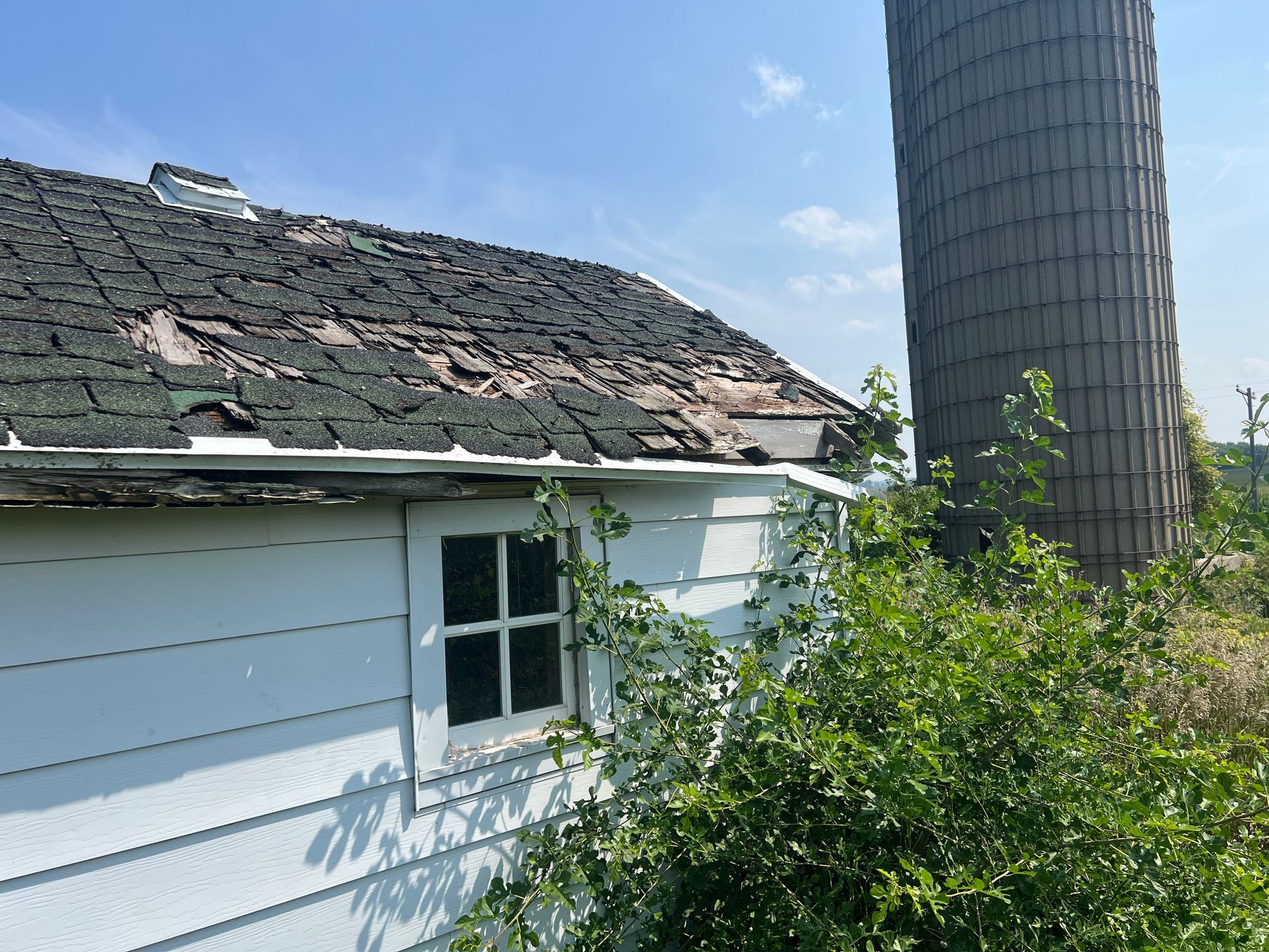 A white building with a damaged, shingled roof sits beside a tall, cylindrical silo under a clear blue sky.