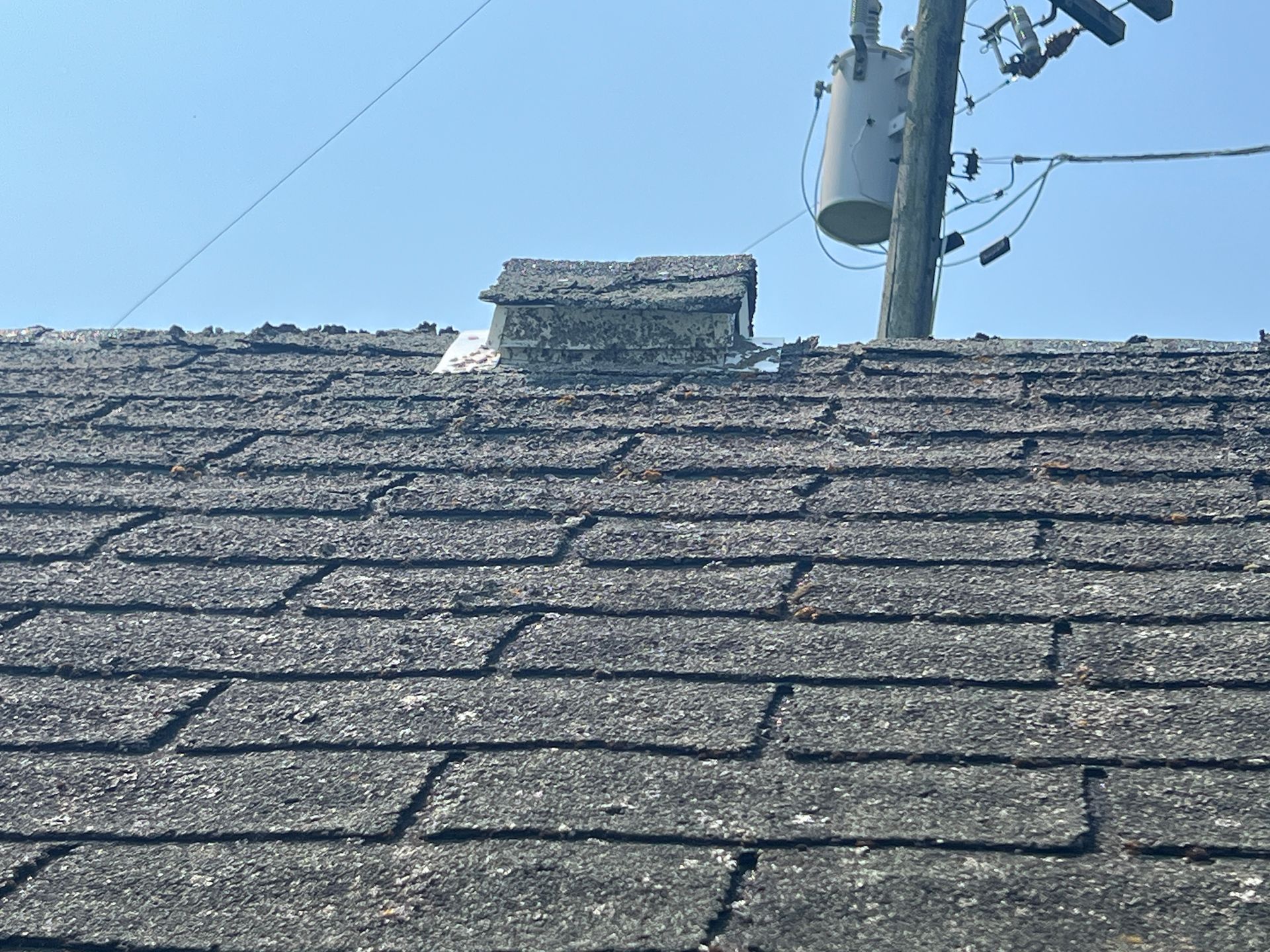 A weathered roof with asphalt shingles, featuring a small, silver-colored ventilation cap, seen against a clear sky.