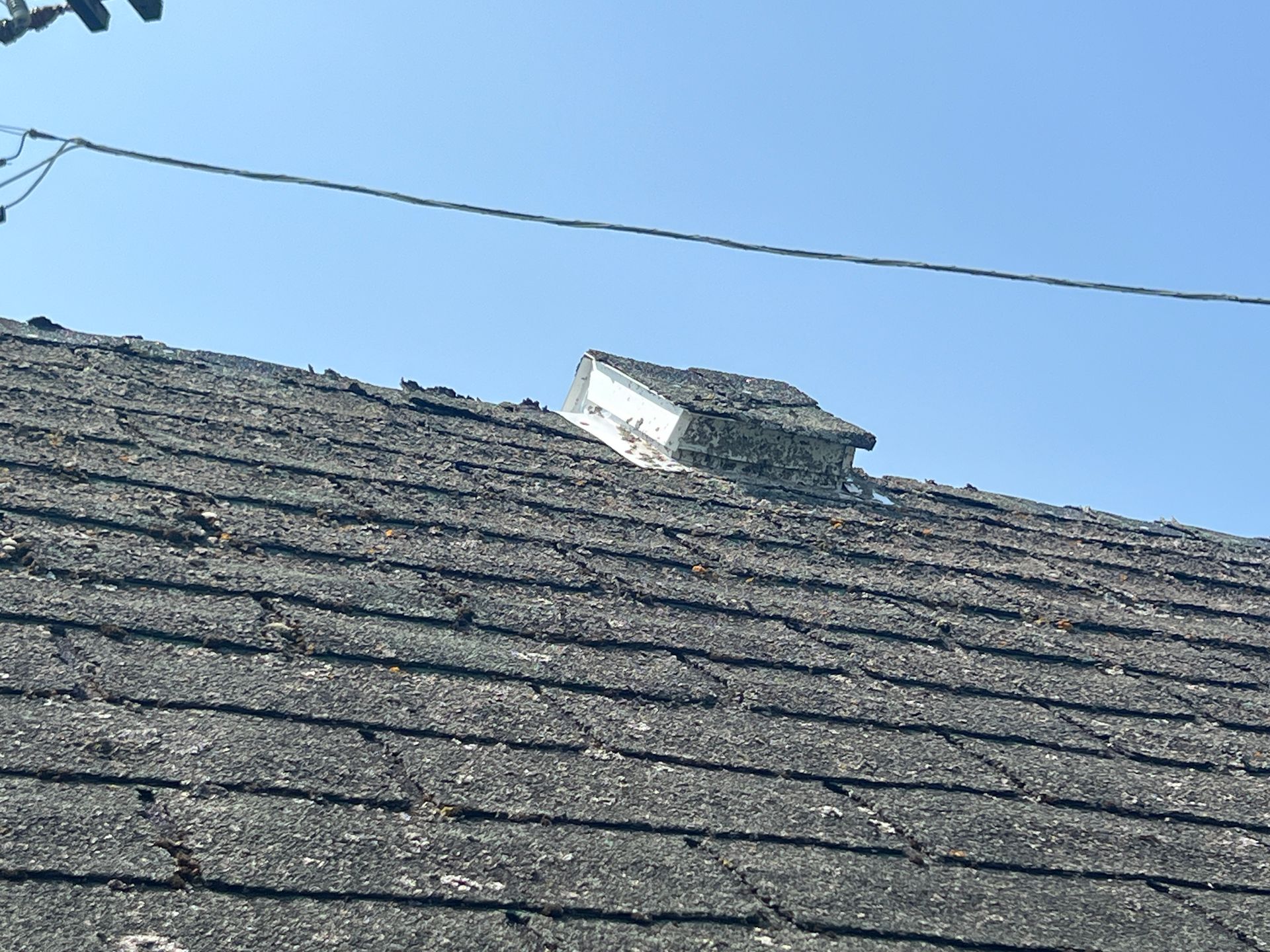 A small, weathered roof vent on an asphalt shingle roof against a clear blue sky.