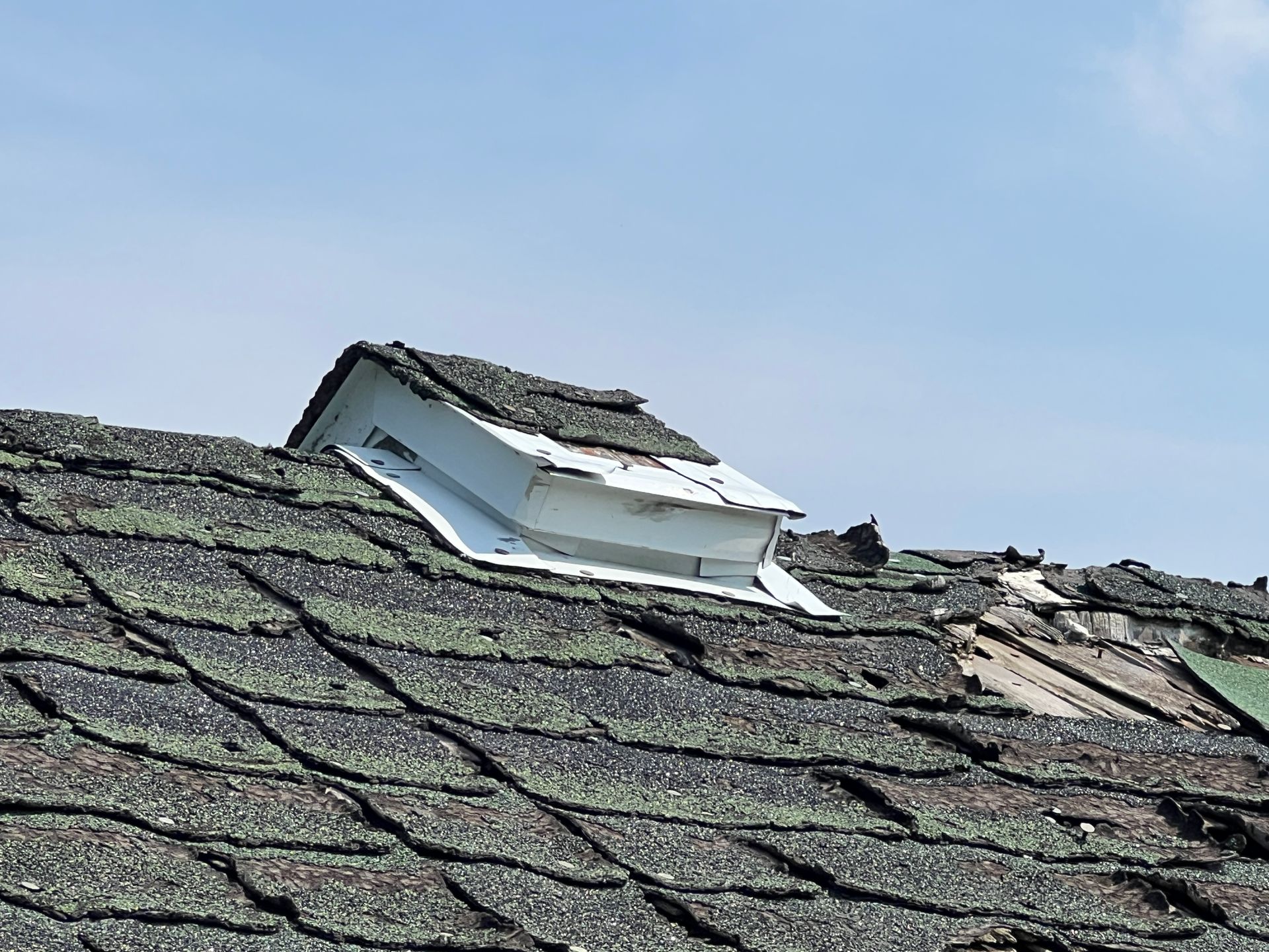 A roof vent with white sides and shingles on top, positioned on a weathered, shingled residential roof.