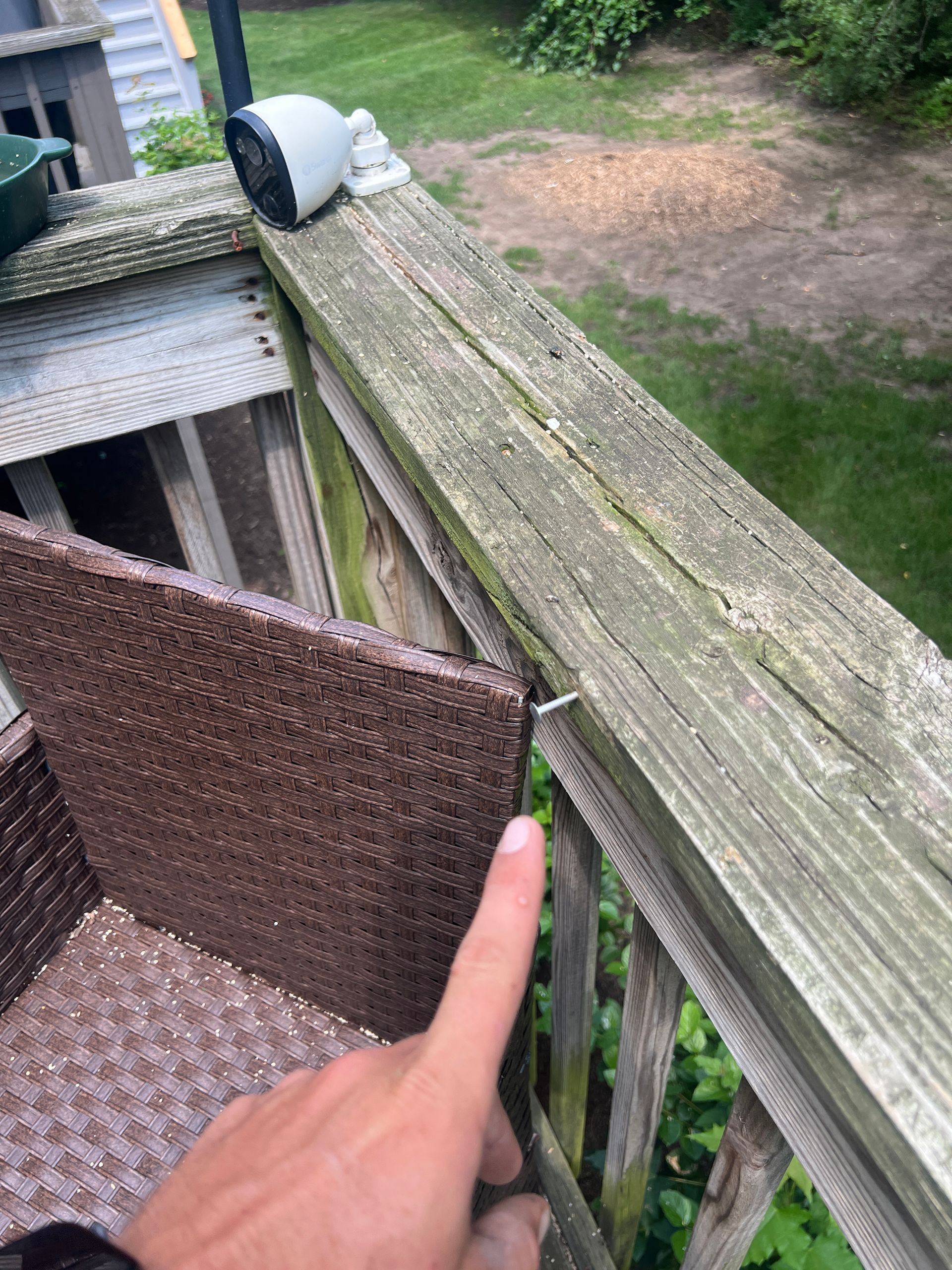 A hand points to a metal nail sticking out of a wooden deck railing near a wicker chair and a security camera.