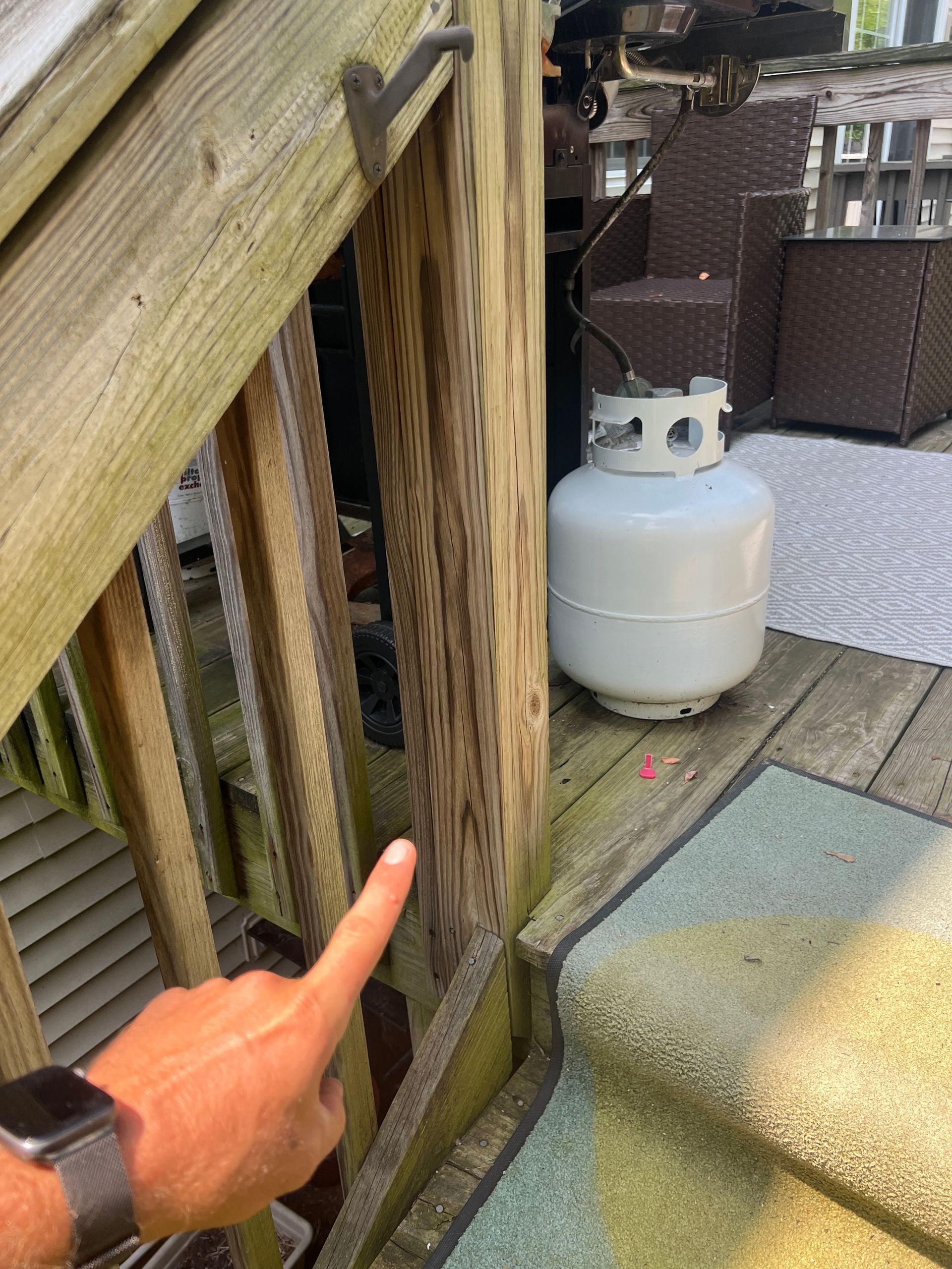 A person's hand points toward a wooden stair railing on a deck next to a gray propane tank.