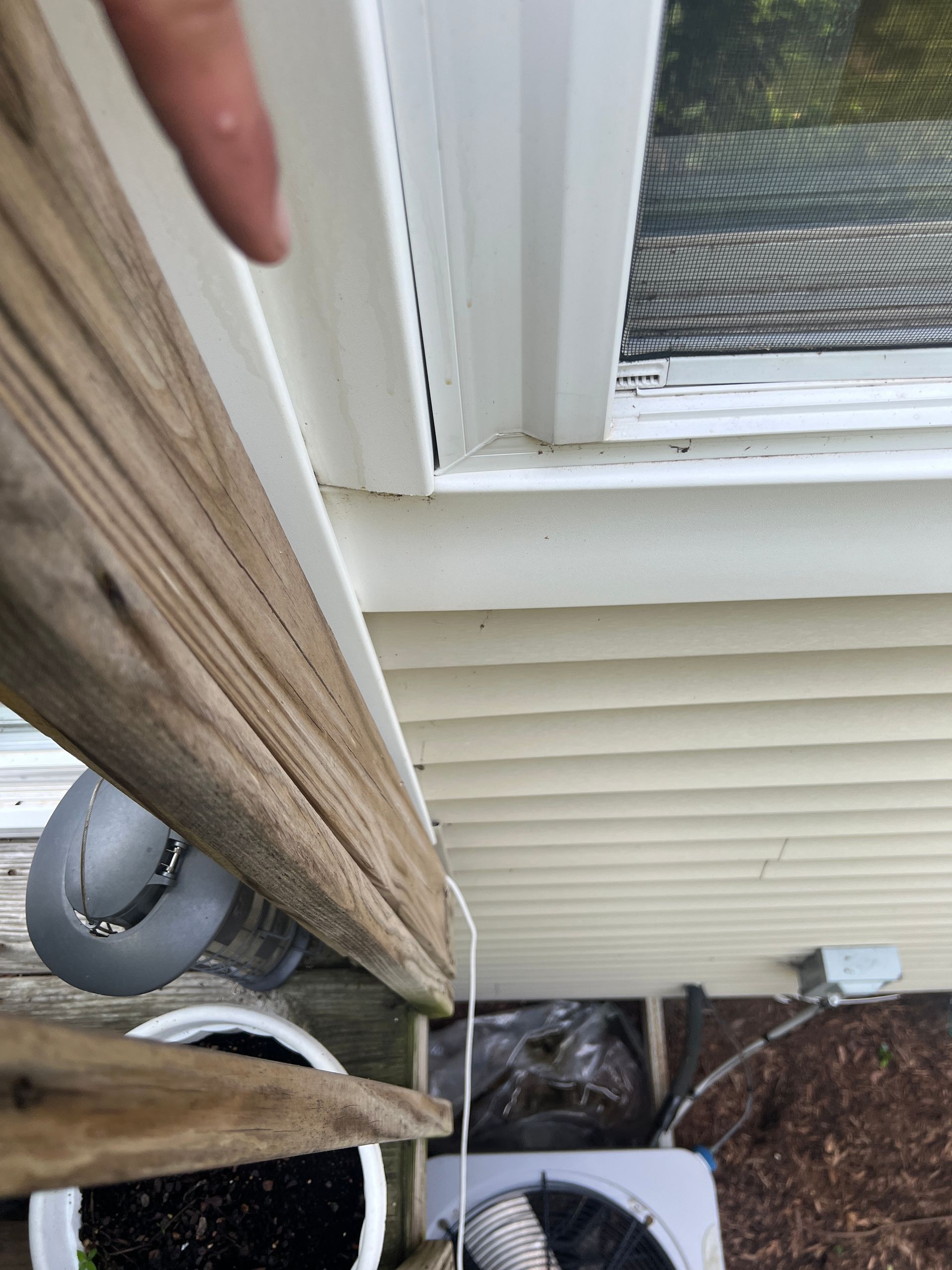 A finger points toward a gap in the white window trim against yellow vinyl siding, near wooden deck stairs and an AC unit.