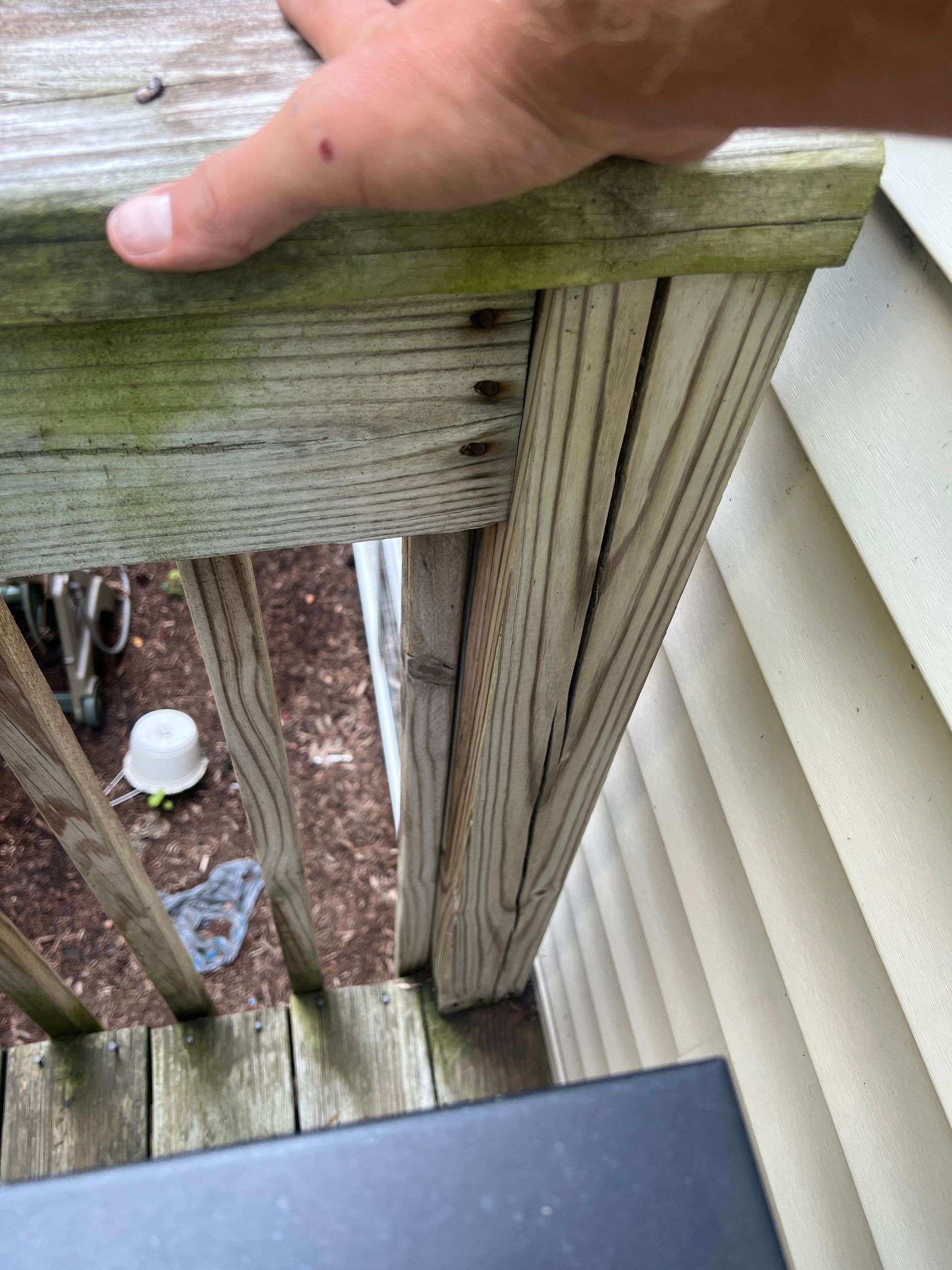 A hand rests on a weathered, outdoor wooden deck railing showing signs of wood splitting and decay near the house siding.