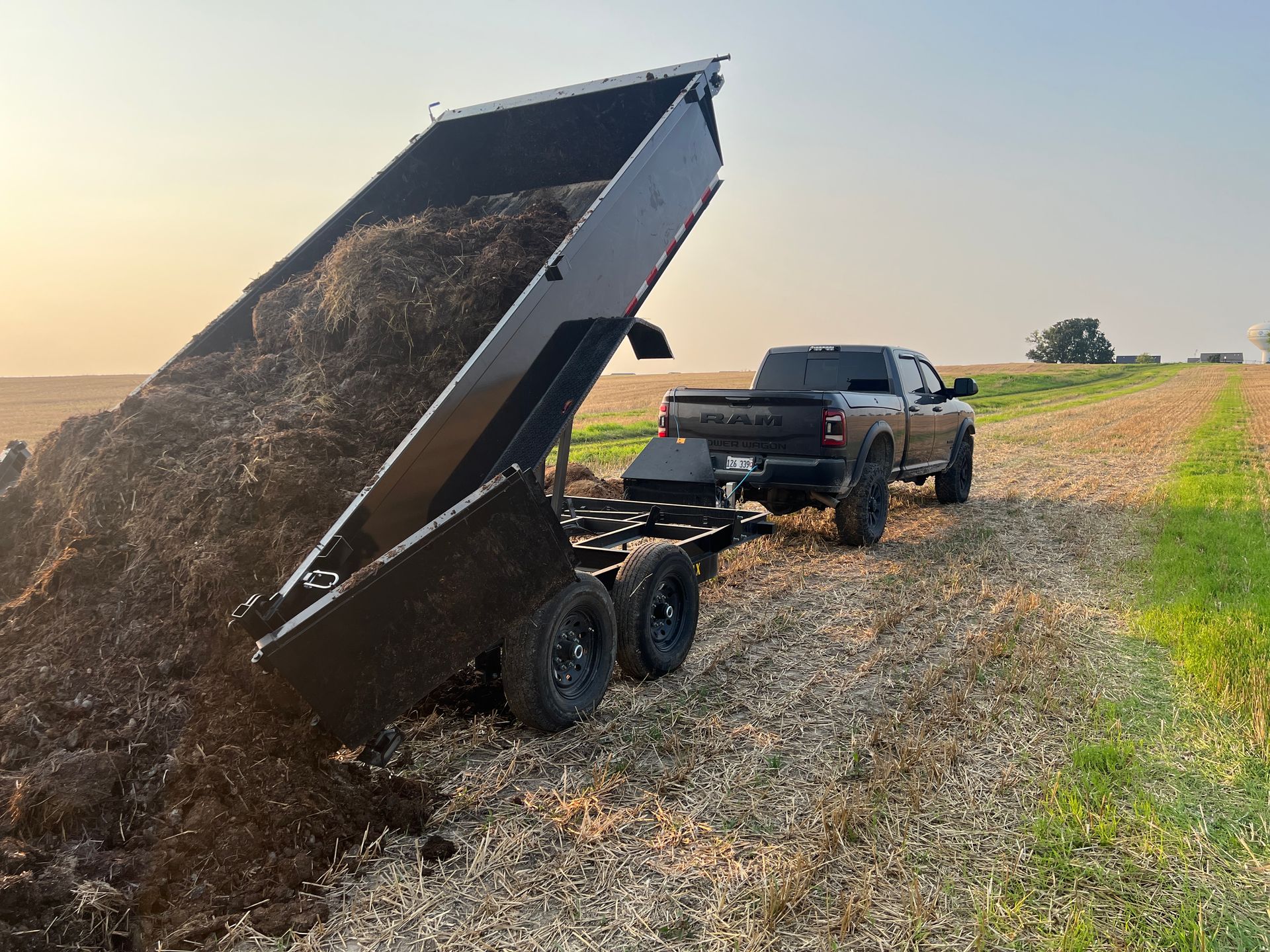 A black pickup truck pulls a dump trailer in a field, with the trailer bed tilted to unload a large pile of dark mulch.